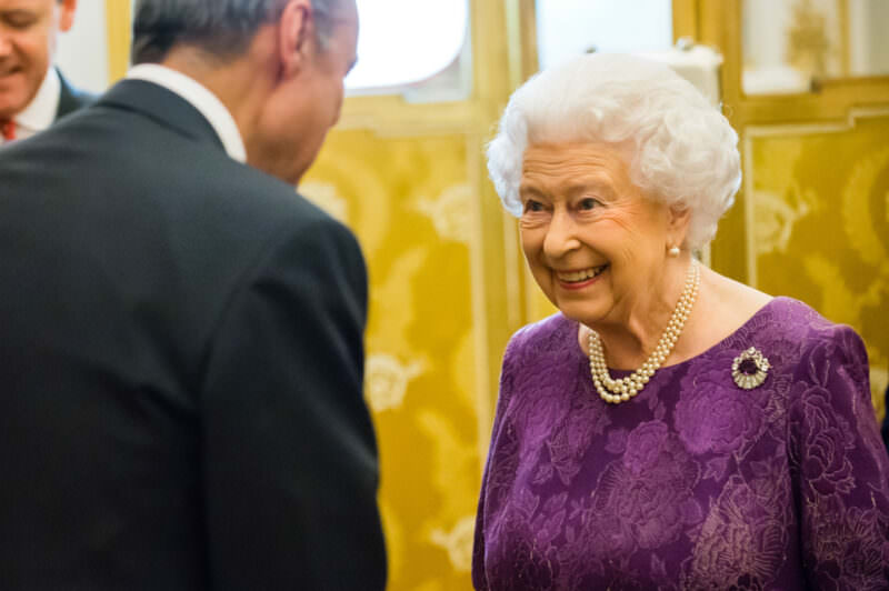 An older woman with white hair, wearing a purple dress and pearl necklace, smiles while speaking to a man in a suit. They are in an elegant room with golden decor. James Gifford-Mead Photography - Event Photographer London