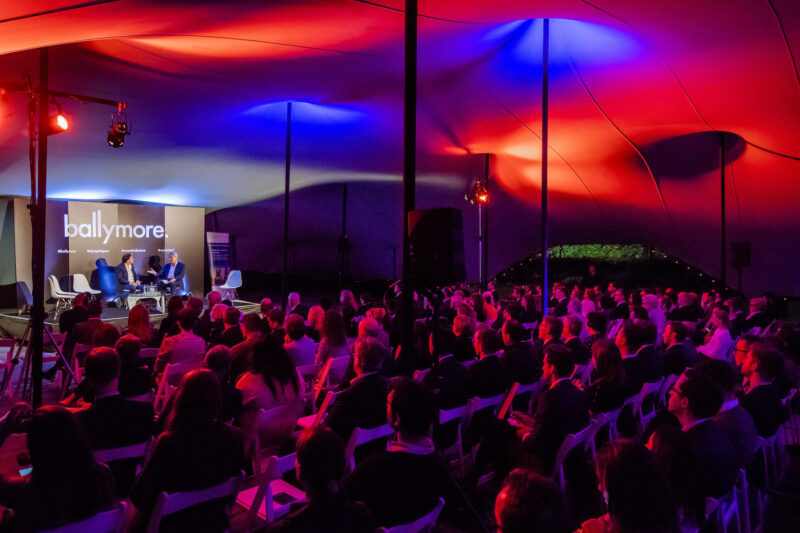 A well-attended indoor event with a large audience seated and listening to a group of speakers on stage. The ceiling is illuminated with vibrant red and blue lights, and a sign reading "ballymore" is visible behind the speakers. James Gifford-Mead Photography - Event Photographer London
