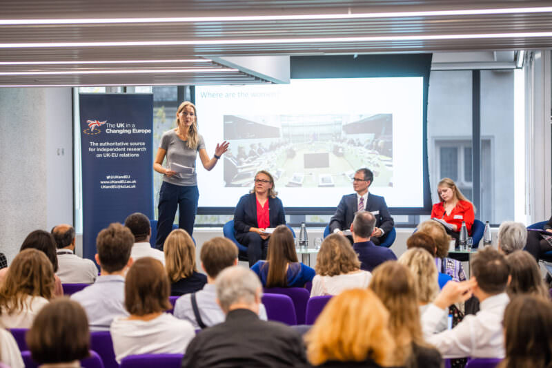 A woman speaks on stage in front of an audience, with three people seated beside her. A screen behind them displays a presentation titled "Where are the women?" and a banner on the left reads "The UK in a Changing Europe. James Gifford-Mead Photography - Event Photographer London
