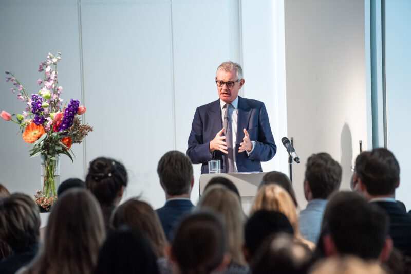 A man in a suit speaks passionately at a podium to a seated audience. A microphone is nearby, and a colorful bouquet of flowers decorates the space. James Gifford-Mead Photography - Event Photographer London
