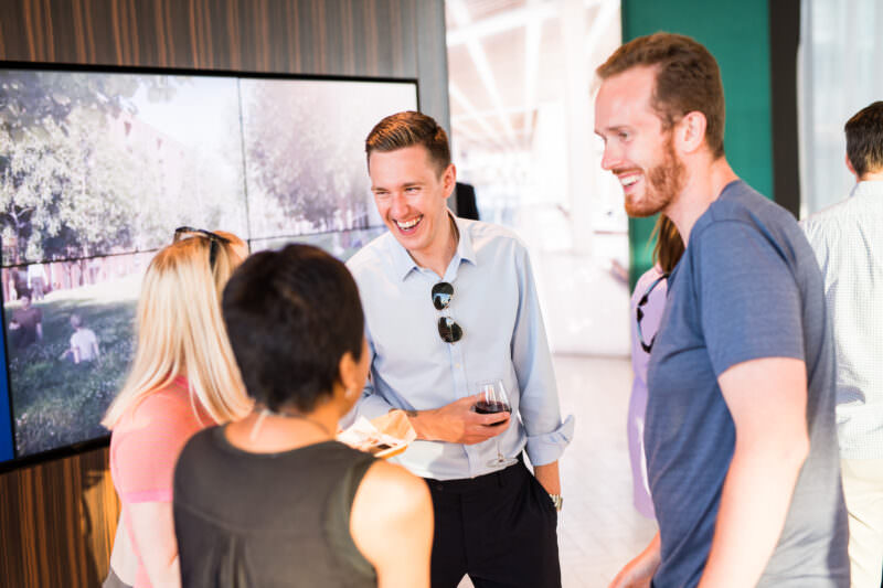 A group of five people stand together in a lively conversation. One man, holding a drink, is laughing. A large display screen is in the background, showing an outdoor scene. The atmosphere appears cheerful and social. James Gifford-Mead Photography - Event Photographer London