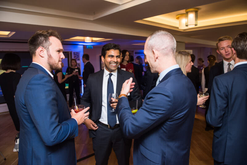 A group of people in formal attire are socializing at an indoor event. Three men in suits stand in the foreground, holding drinks and smiling. More attendees are visible in the background under warm lighting. James Gifford-Mead Photography - Event Photographer London