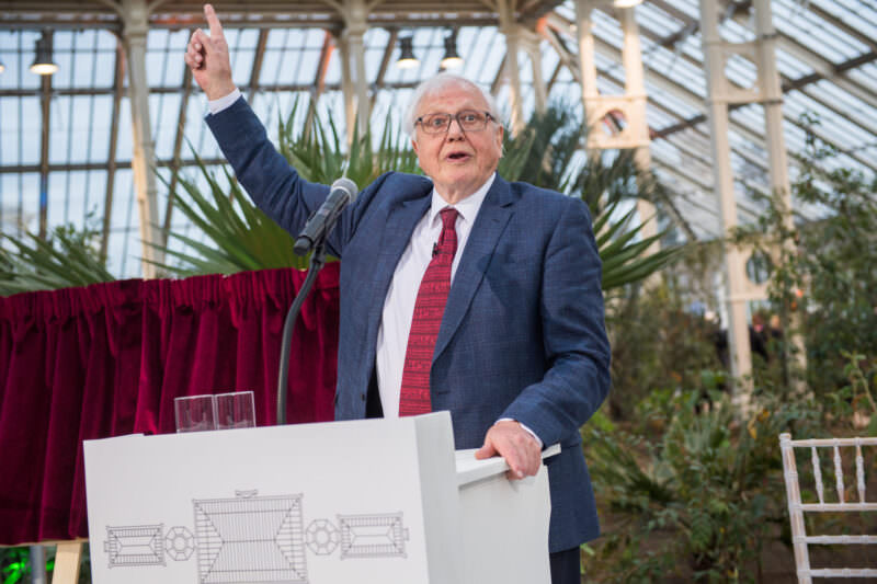 An elderly man in a suit and red tie stands at a podium, speaking animatedly with one arm raised. He is inside a greenhouse with large plants and glass panels visible in the background. A red curtain is partially visible to his left. James Gifford-Mead Photography - Event Photographer London