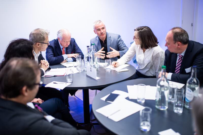 Seven people sit around a circular table in a discussion, with notebooks and bottles of water. A man gestures with his hands while speaking, and others listen attentively. The setting is a brightly lit conference room. James Gifford-Mead Photography - Event Photographer London