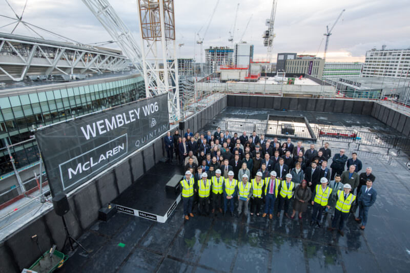 A large group of construction workers and professionals wearing safety vests and hard hats stand on a rooftop platform. Behind them, a tall crane and multiple buildings are visible. A banner reads "Wembley W06" with "McLaren" and "Quintain" logos. James Gifford-Mead Photography - Event Photographer London