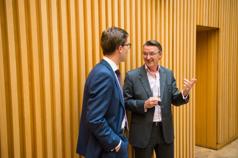 Two men in business attire stand talking in front of a wooden panel wall. The man on the left wears a blue suit and glasses, facing the man on the right, who wears a gray suit and gestures with his hand while holding a glass. James Gifford-Mead Photography - Event Photographer London