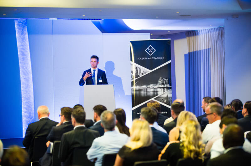A man in a suit is speaking at a podium in front of an audience in a conference room. There is a banner with the text "Mason Alexander" beside him. Attendees are seated, facing the speaker. James Gifford-Mead Photography - Event Photographer London