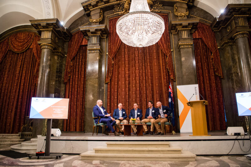 A panel of six people is seated on a stage in an ornate room with tall columns and a large chandelier. They are engaged in a discussion. A podium and a screen displaying graphics are on either side of the stage. James Gifford-Mead Photography - Event Photographer London