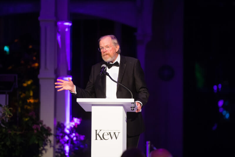 A man in a tuxedo speaks at a lectern with "Kew" written on it. The background is dimly lit, featuring purple lights and foliage. James Gifford-Mead Photography - Event Photographer London