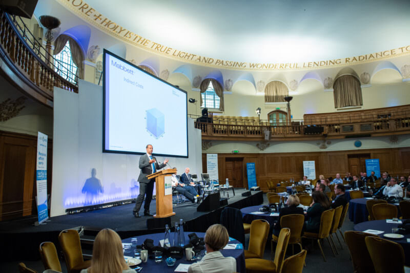 A man is presenting on stage in a large conference hall with a high ceiling and balcony. Attendees sit at tables with laptops and papers. A screen displays a slide titled "Notebook ODM." Busts and a quote adorn the walls. James Gifford-Mead Photography - Event Photographer London