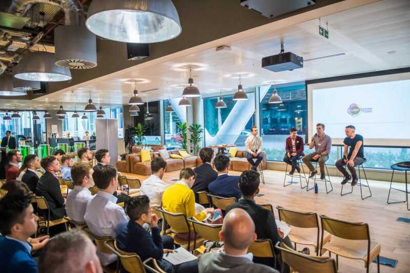 People seated in rows facing a stage where four panelists sit on stools. The room has large windows, overhead lights, and plants. Audience members are listening attentively, and a projector displays a logo on the wall. James Gifford-Mead Photography - Event Photographer London