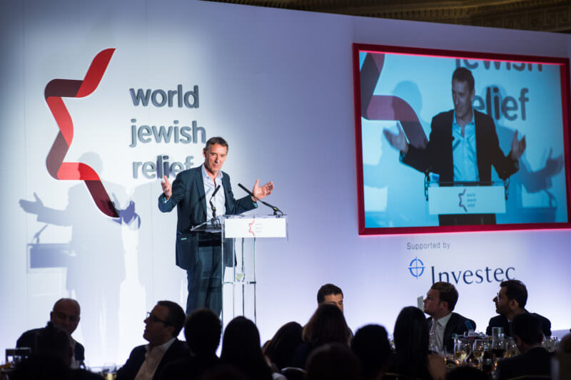 A speaker stands at a podium on a stage, addressing an audience at an event. The backdrop features the text "world jewish relief," and a screen displays the speaker. People are seated at tables in the foreground. James Gifford-Mead Photography - Event Photographer London