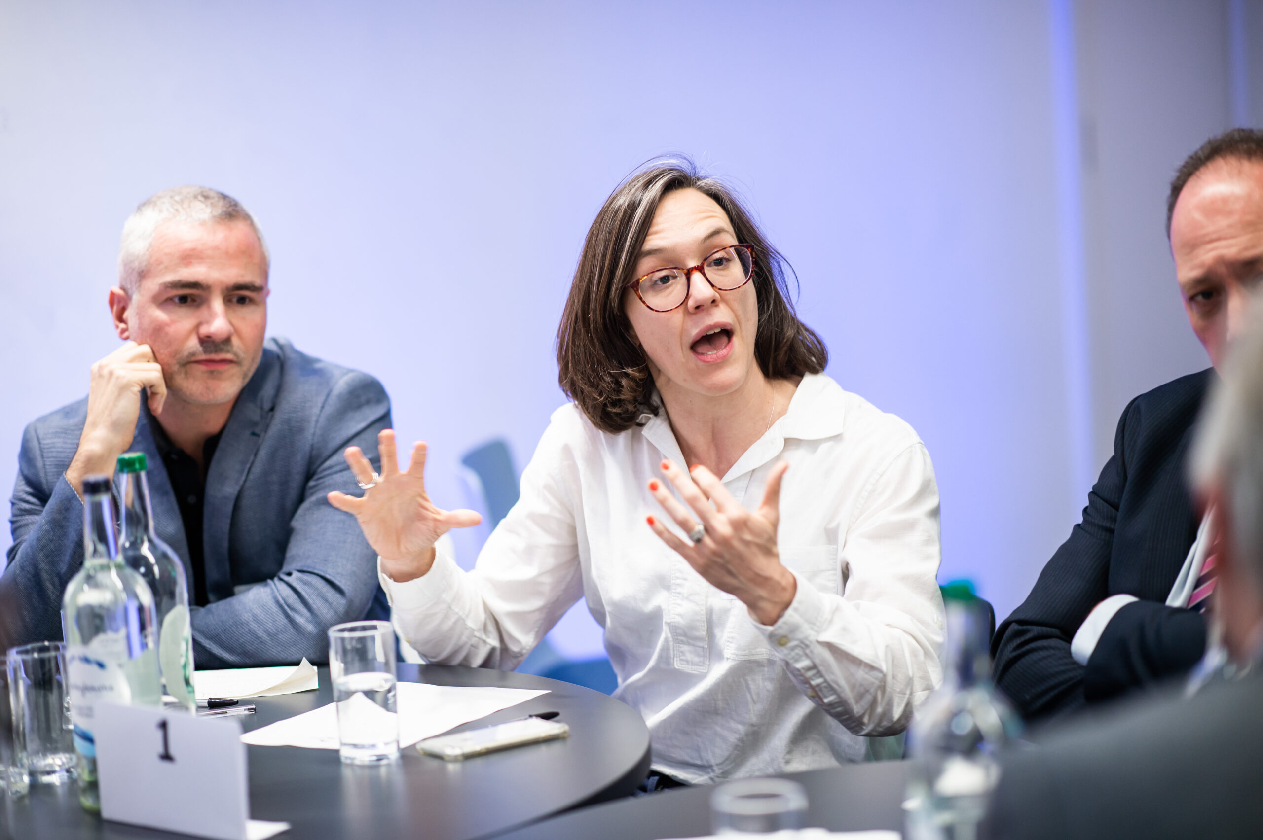 A woman with glasses and a white shirt speaks animatedly, gesturing with her hands during a discussion at a round table with two men and water bottles. James Gifford-Mead Photography - Event Photographer London