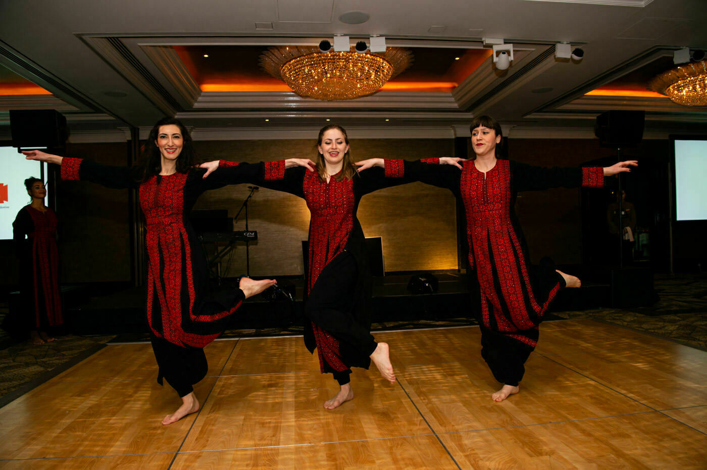 Three women in black and red traditional dresses perform a synchronized dance on a wooden floor in a brightly lit room with chandeliers. Their arms are elegantly outstretched, balancing on one leg. James Gifford-Mead Photography - Event Photographer London