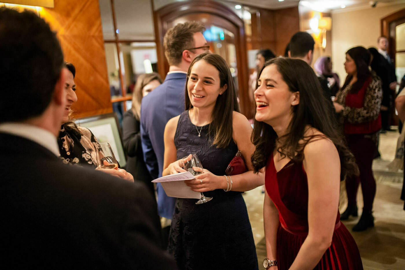 A group of people are engaged in conversation at a social event. Two women in the foreground are smiling and holding drinks. The setting appears to be a warmly lit indoor venue with other people mingling in the background. James Gifford-Mead Photography - Event Photographer London