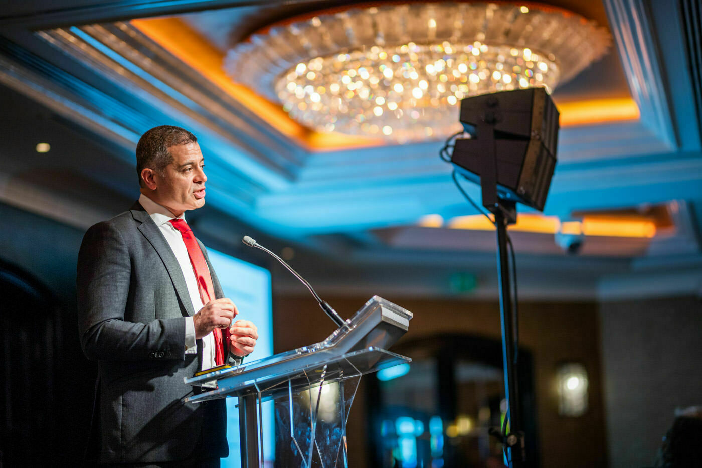 A person in a suit and red tie speaks at a podium with a microphone in a well-lit, elegant room. A chandelier and a screen are visible in the background. James Gifford-Mead Photography - Event Photographer London
