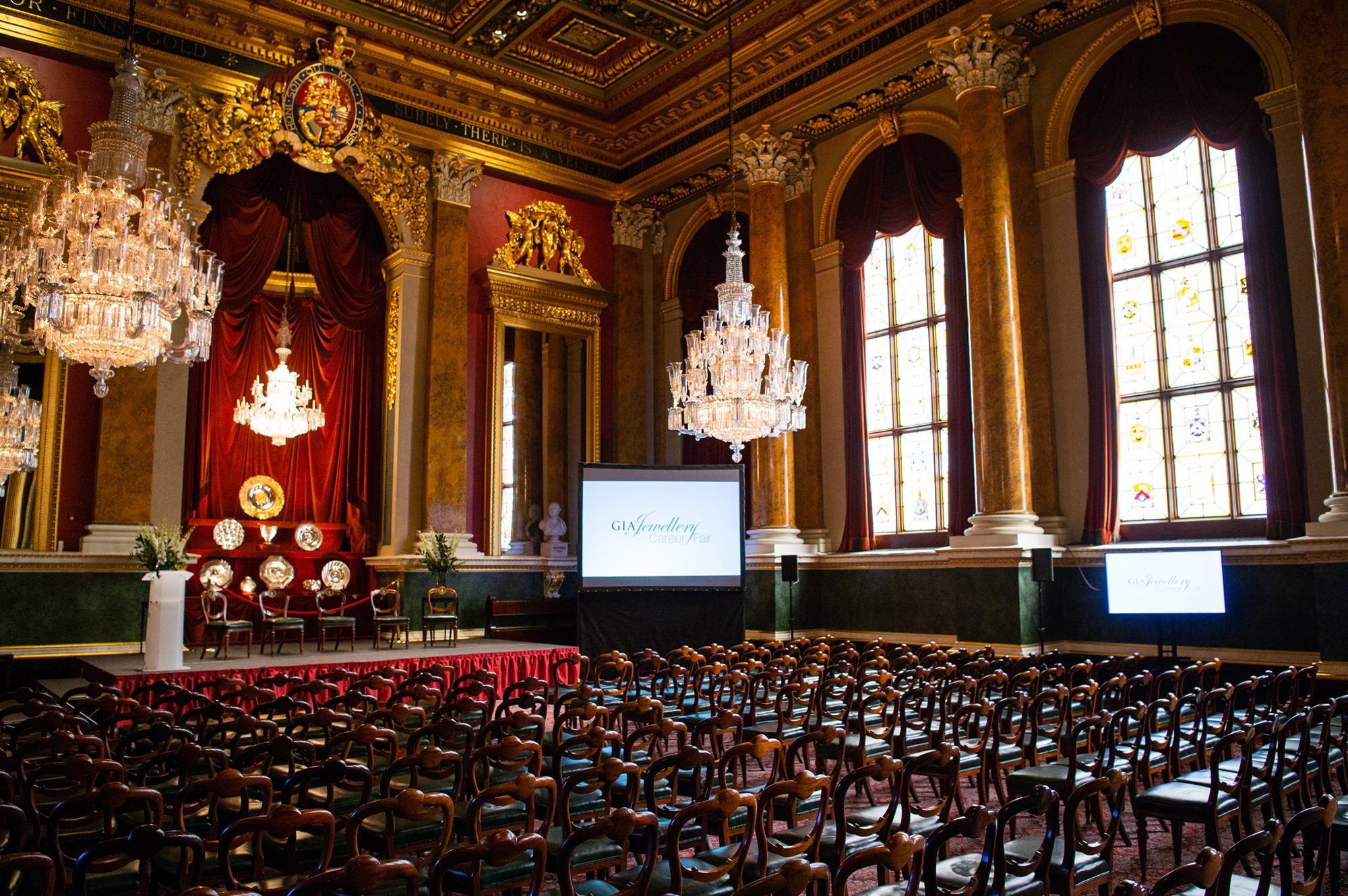 An elegant, ornately decorated hall with rows of empty wooden chairs facing a stage adorned with red drapes, chandeliers, and large windows. Two screens display "Gathering" next to potted plants on stage. James Gifford-Mead Photography - Event Photographer London
