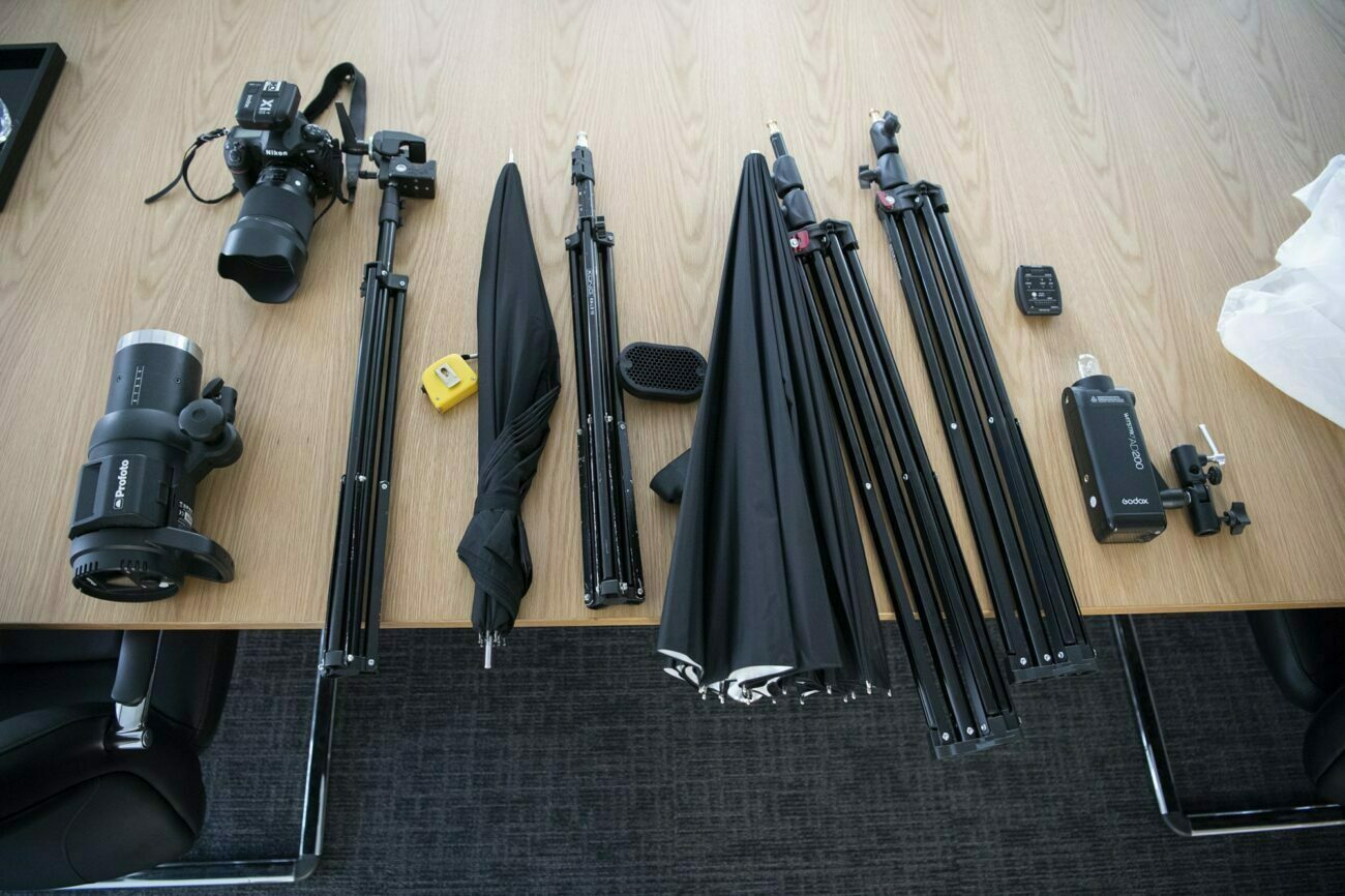 Photography equipment spread on a wooden table, including a camera, lenses, tripods, light stands, umbrellas, a tape measure, and a remote trigger. The items are neatly arranged in rows, suggesting preparation for a photoshoot. James Gifford-Mead Photography - Event Photographer London