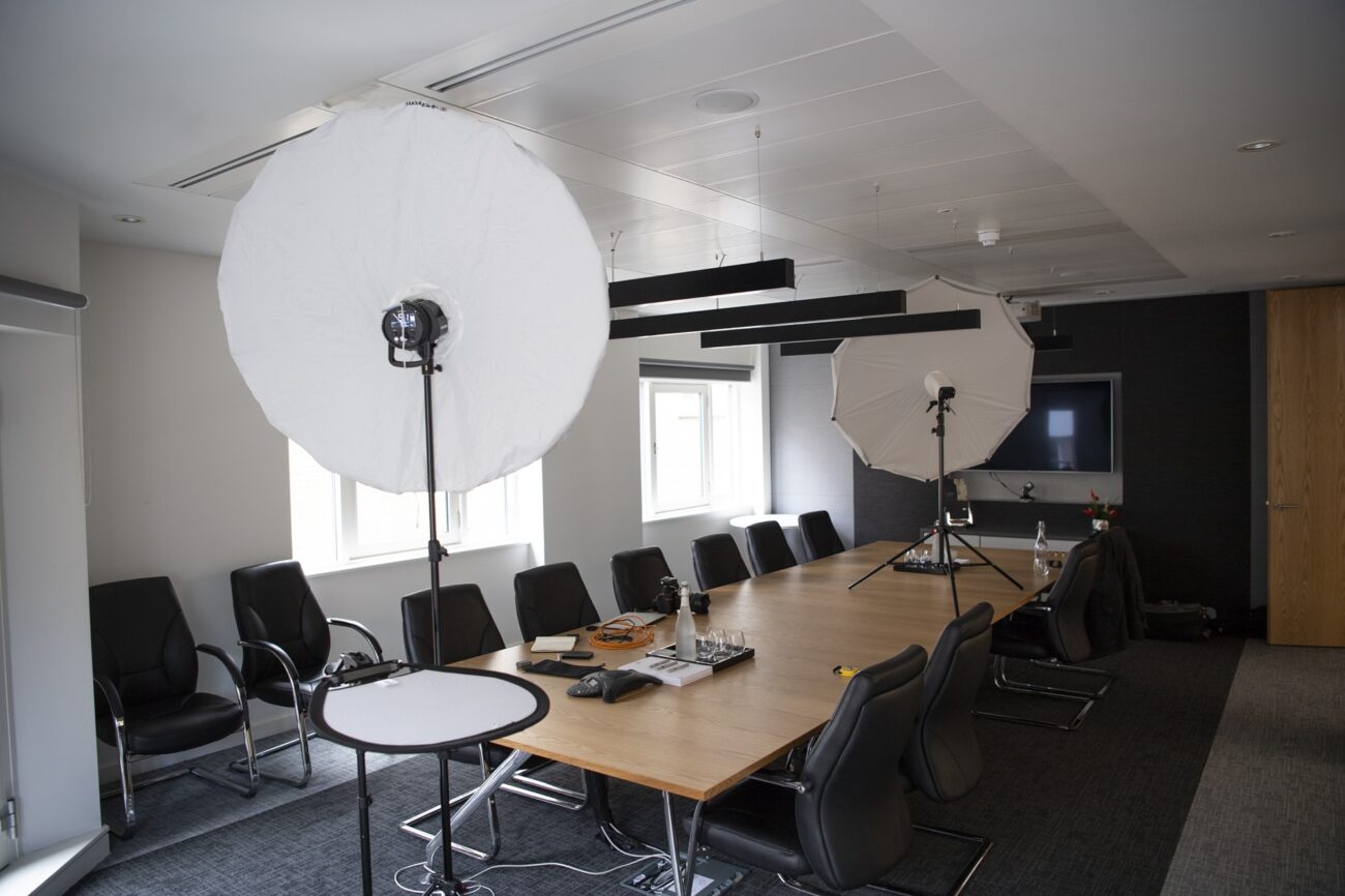 A conference room setup for a photo shoot with large softbox lights around a long wooden table surrounded by black chairs. Cables, a laptop, and camera equipment are placed on the table. A flat-screen TV is mounted on the wall. James Gifford-Mead Photography - Event Photographer London