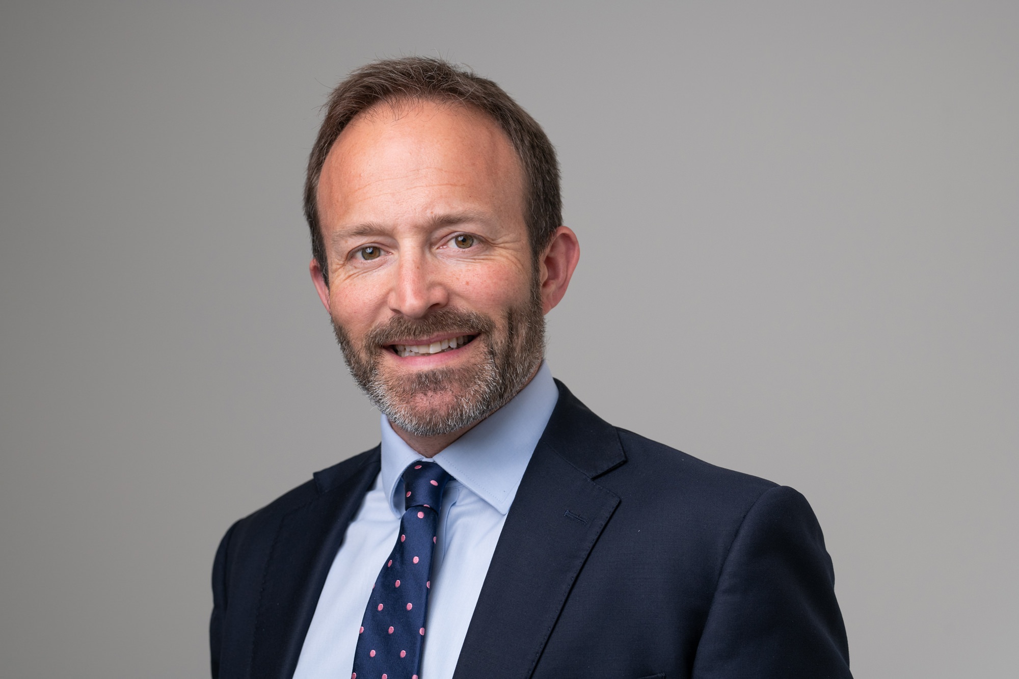 Man in a dark suit and a polka dot tie smiling against a neutral background. James Gifford-Mead Photography - Event Photographer London