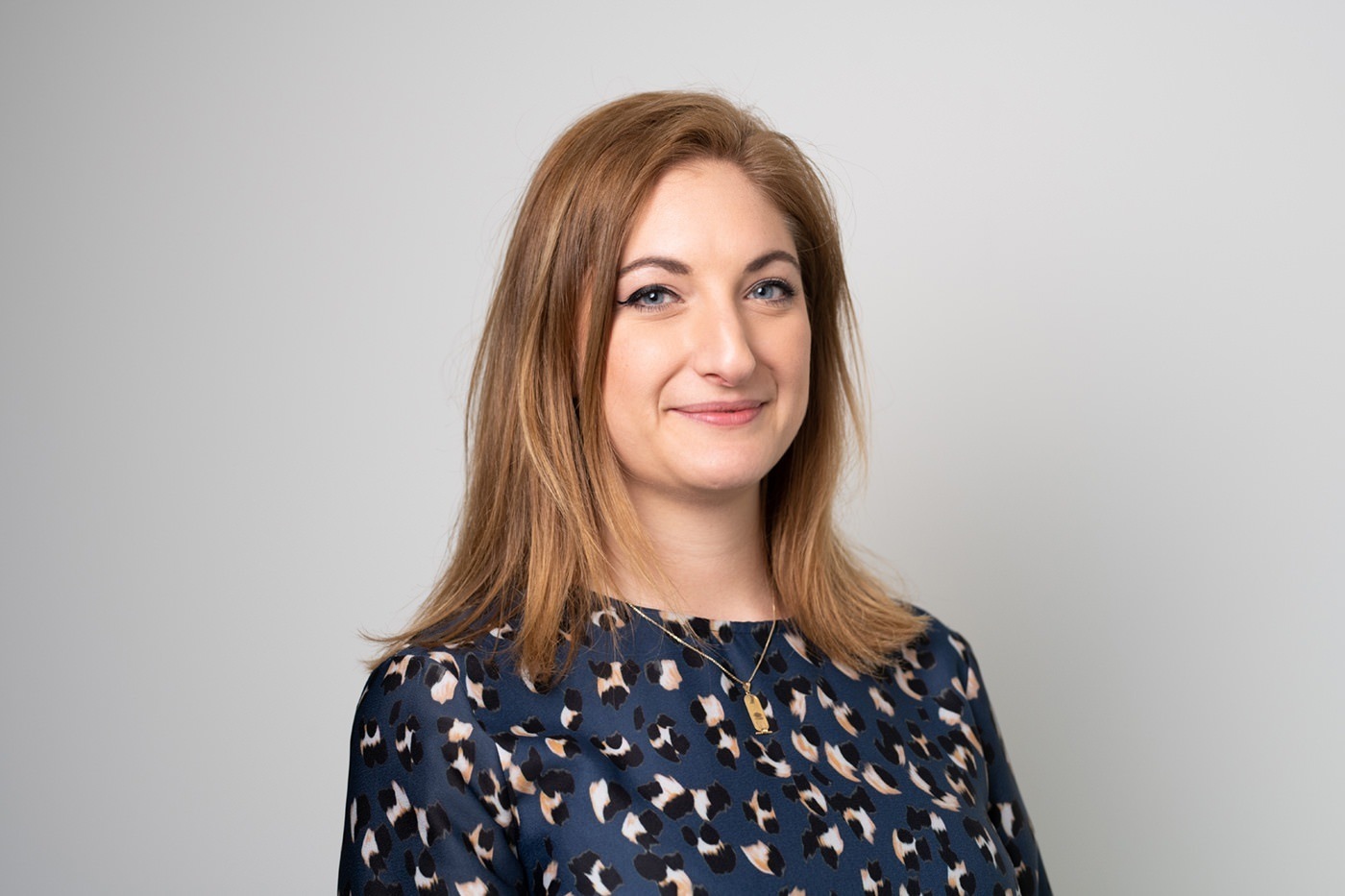 Woman with long light brown hair wearing a leopard print blouse, standing against a plain light gray background, smiling slightly while looking at the camera. James Gifford-Mead Photography - Event Photographer London