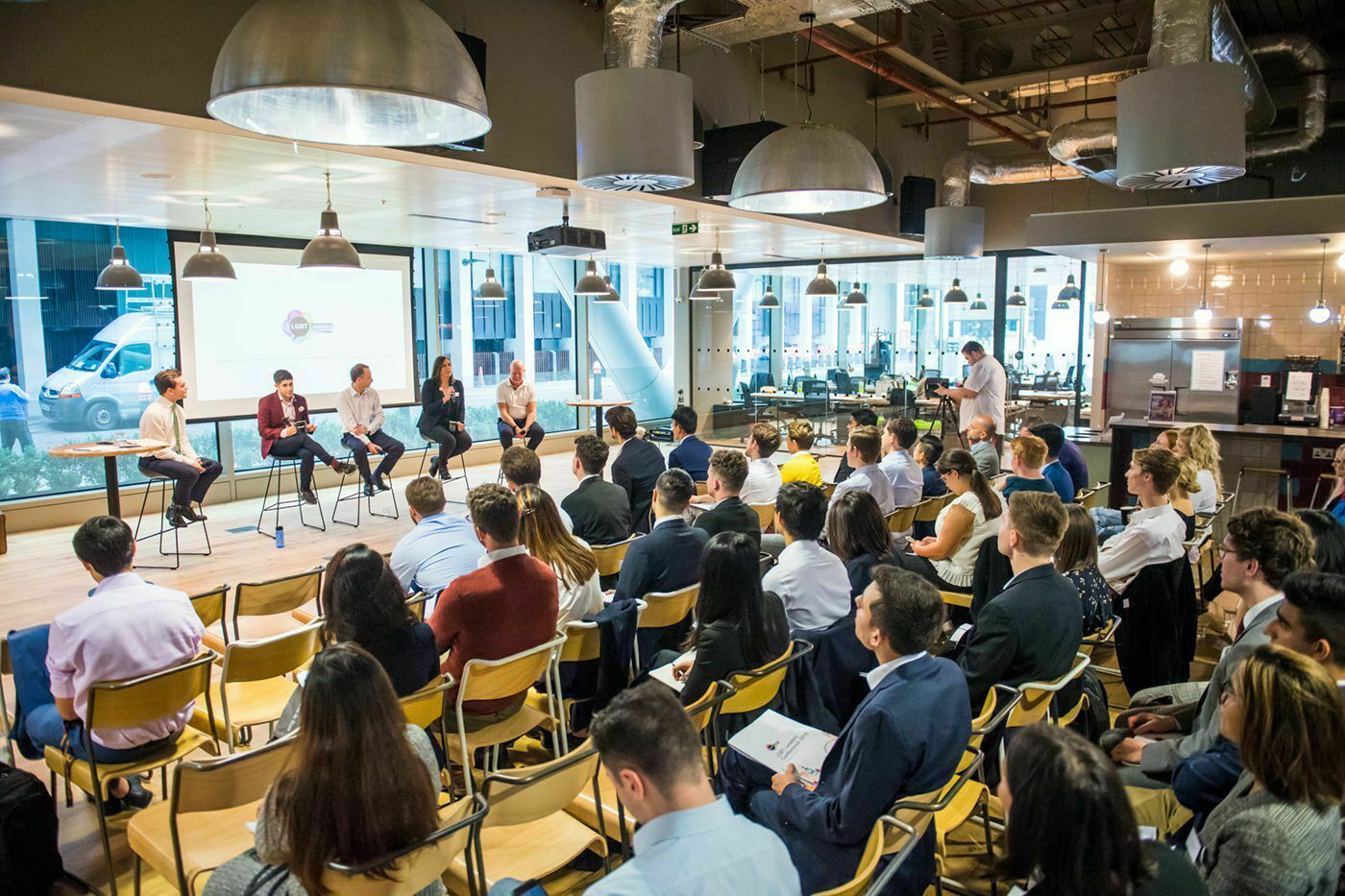 A diverse group of people sit in a modern conference room facing a panel of five speakers on stage. Large screens and industrial lighting are overhead. The audience is attentively listening. James Gifford-Mead Photography - Event Photographer London
