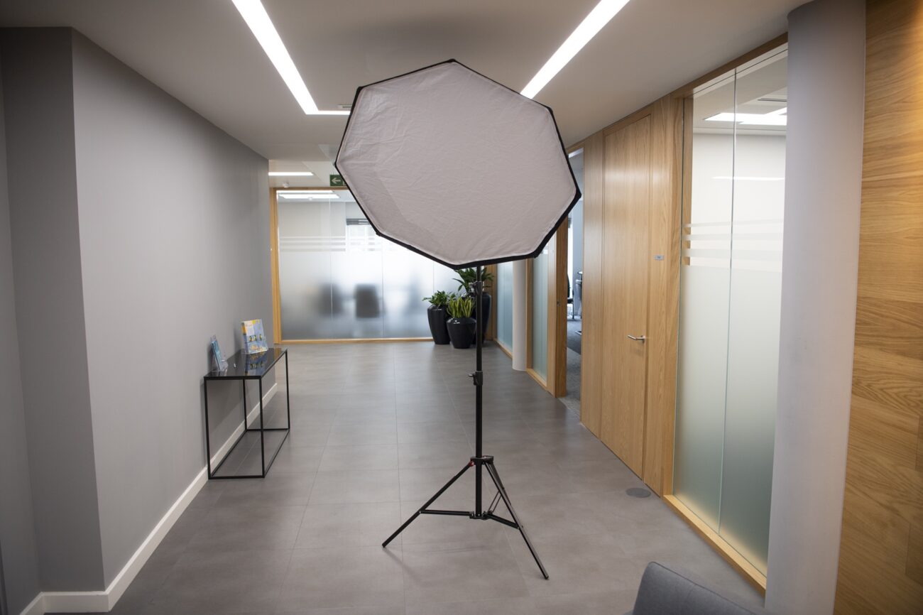 A photography softbox light is set up in a modern office hallway with gray walls and a tiled floor. The corridor is lined with wooden doors and glass panels, and a small table holds brochures near potted plants. James Gifford-Mead Photography - Event Photographer London