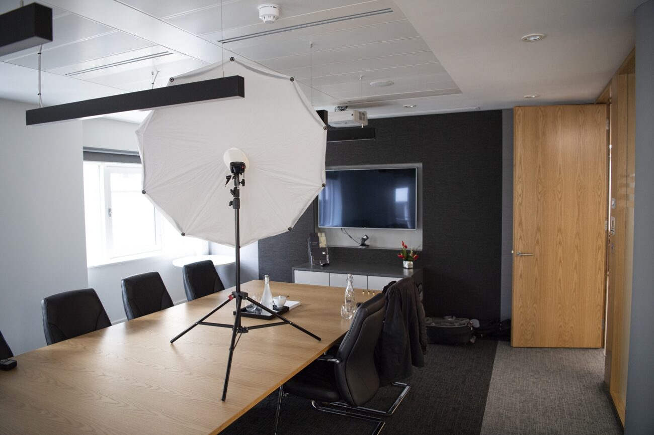 A conference room with a wooden table, black chairs, and a large TV on a dark wall. A photography umbrella and lighting equipment are set up near the window. There is a wooden door on the right and a few items on the table, like water bottles. James Gifford-Mead Photography - Event Photographer London