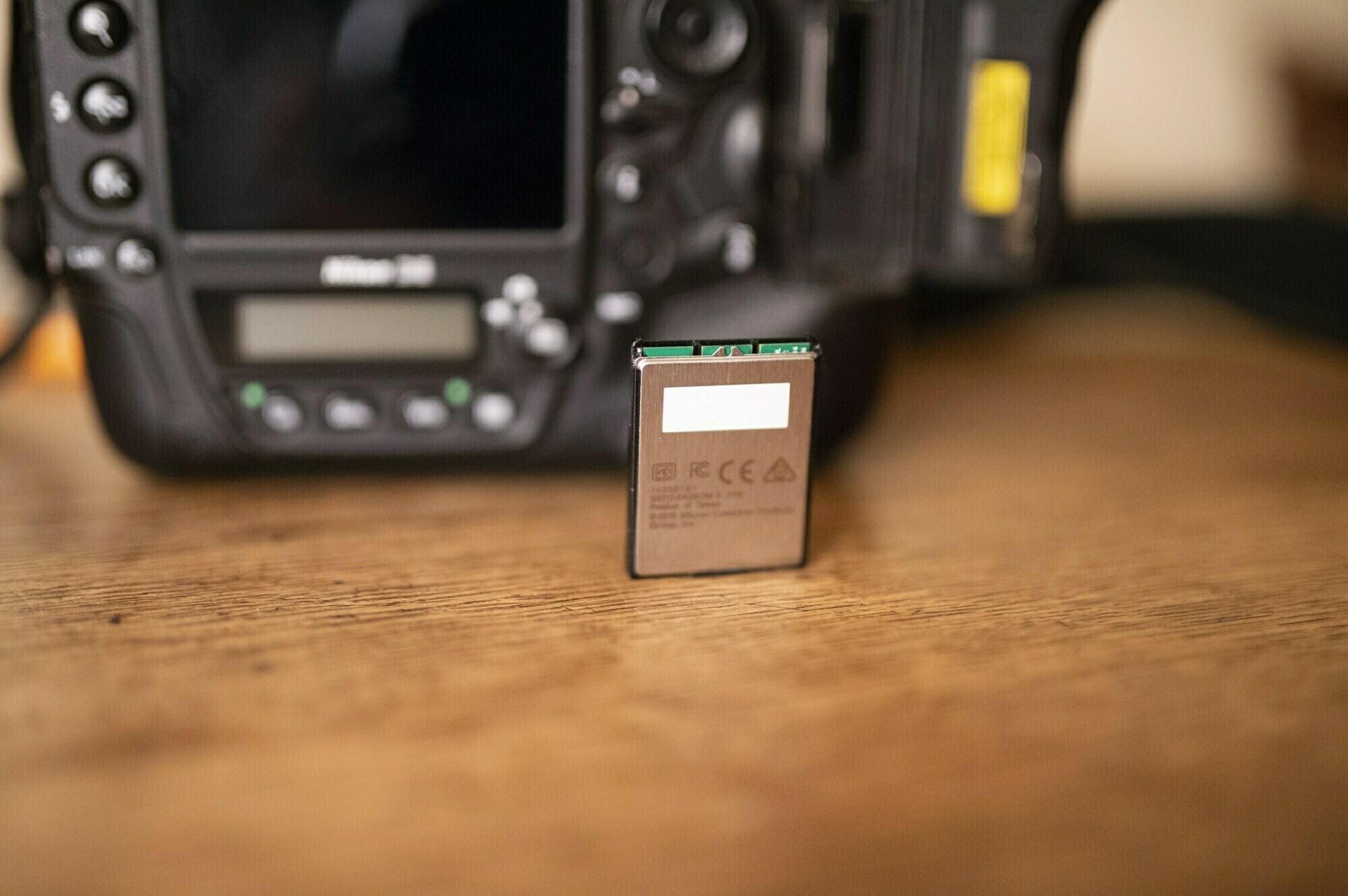A close-up of a camera battery standing upright on a wooden surface, with a blurred DSLR camera in the background. James Gifford-Mead Photography - Event Photographer London