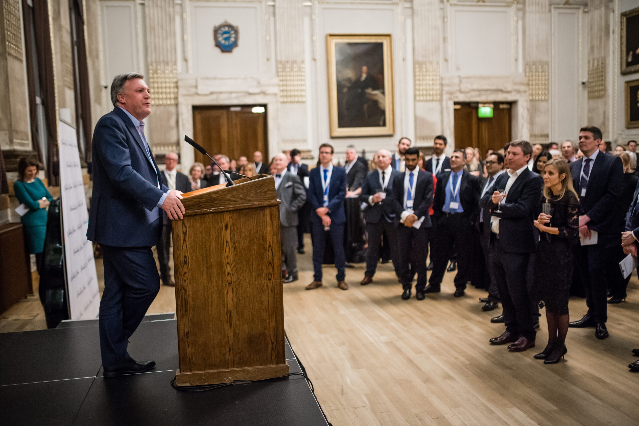 A man in a suit speaks at a wooden podium to a crowd in a formal room with ornate details and paintings. The audience, wearing business attire, listens attentively while standing. James Gifford-Mead Photography - Event Photographer London