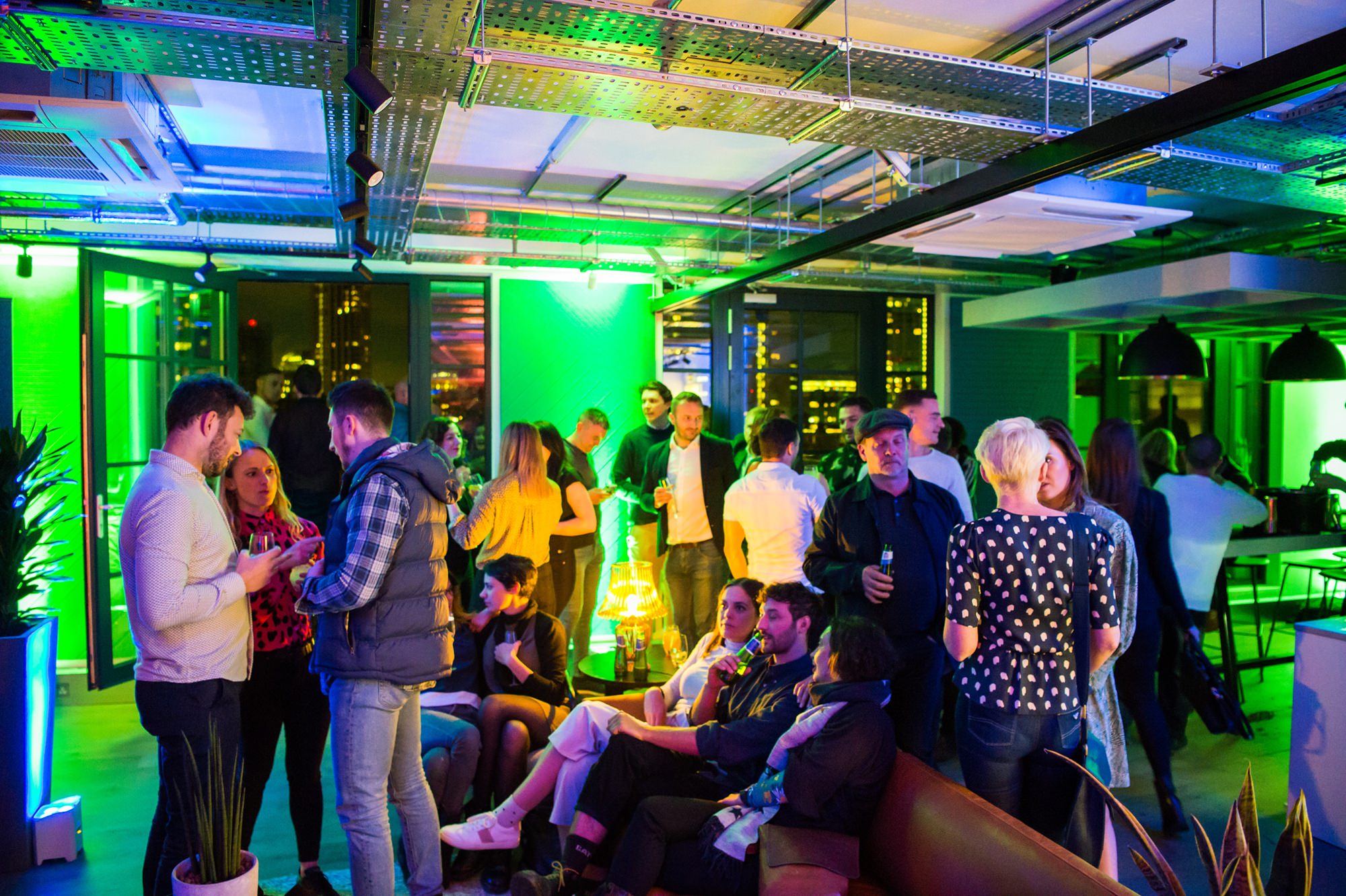A lively indoor party scene with people socializing in a modern space with colorful lights. Some are standing and talking, while others sit on a couch. The room features large windows, plants, and a view of city lights in the background. James Gifford-Mead Photography - Event Photographer London