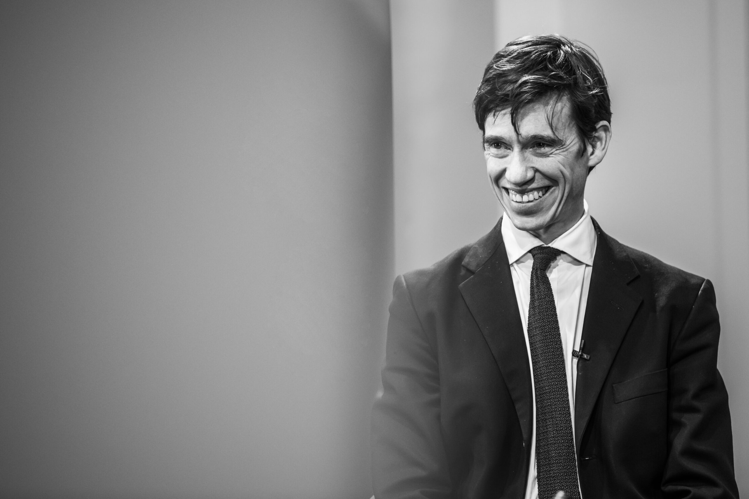 A man in a suit and tie is smiling while sitting in a chair. The image is in black and white, and he is positioned to the right with a blurred background on the left. James Gifford-Mead Photography - Event Photographer London