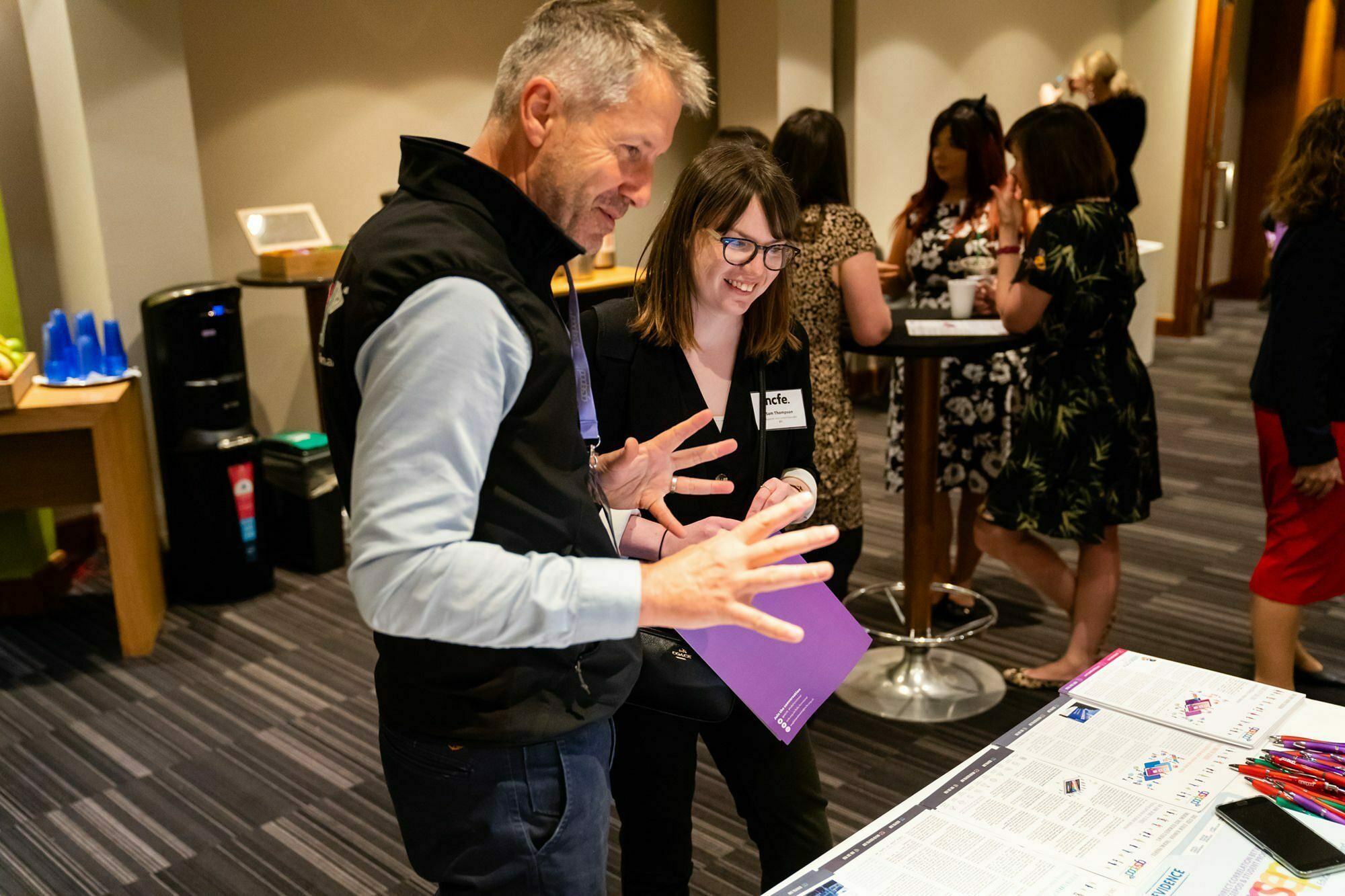 A man and woman engaged in conversation by a table with papers and maps in a conference room. Other people are talking in the background. The room has tables with drinks and office supplies. James Gifford-Mead Photography - Event Photographer London