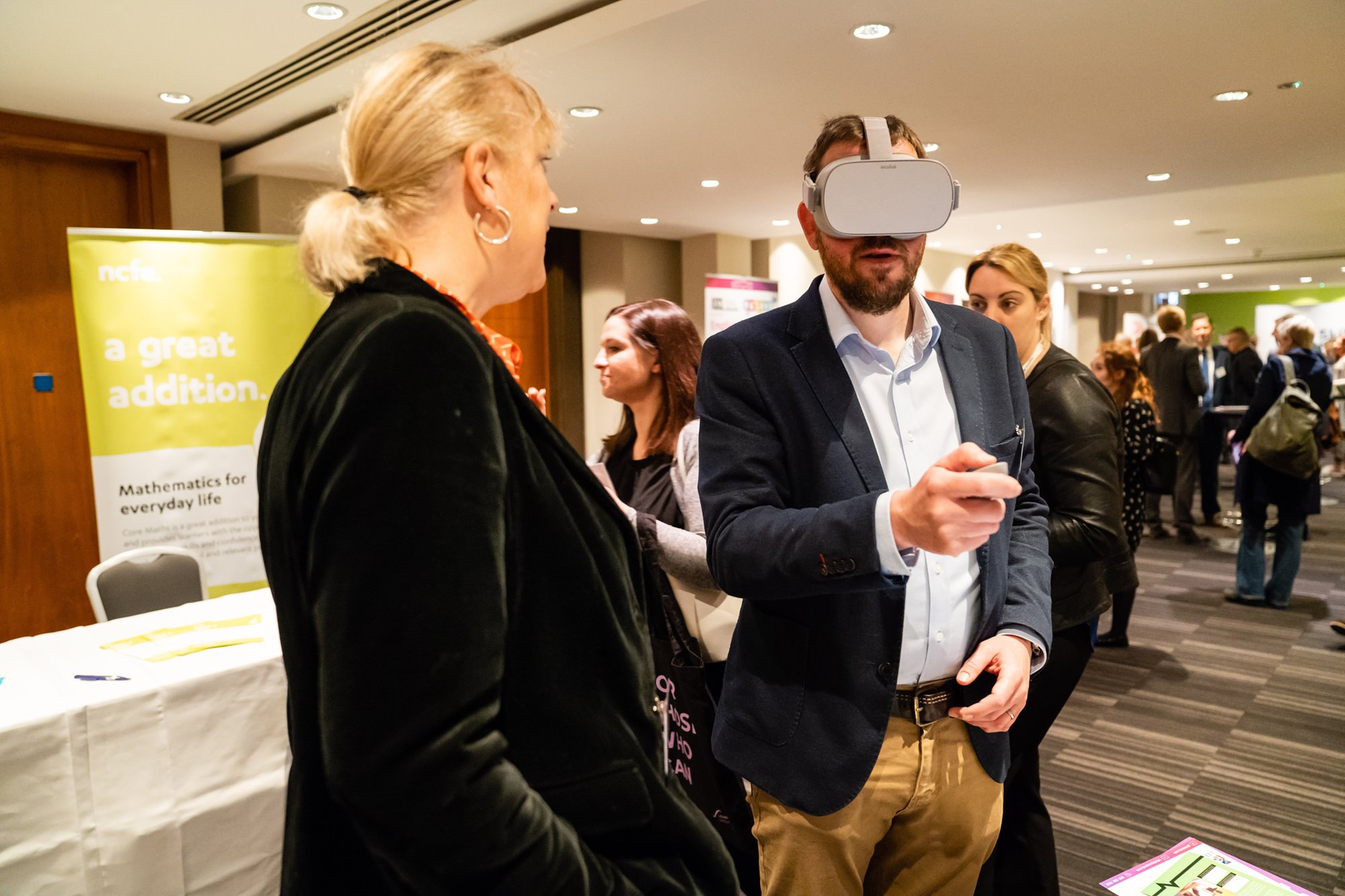 A man wearing a VR headset and holding a controller interacts with a display at an indoor event. A woman stands nearby watching. Other attendees are in the background, and a banner about mathematics is partially visible. James Gifford-Mead Photography - Event Photographer London