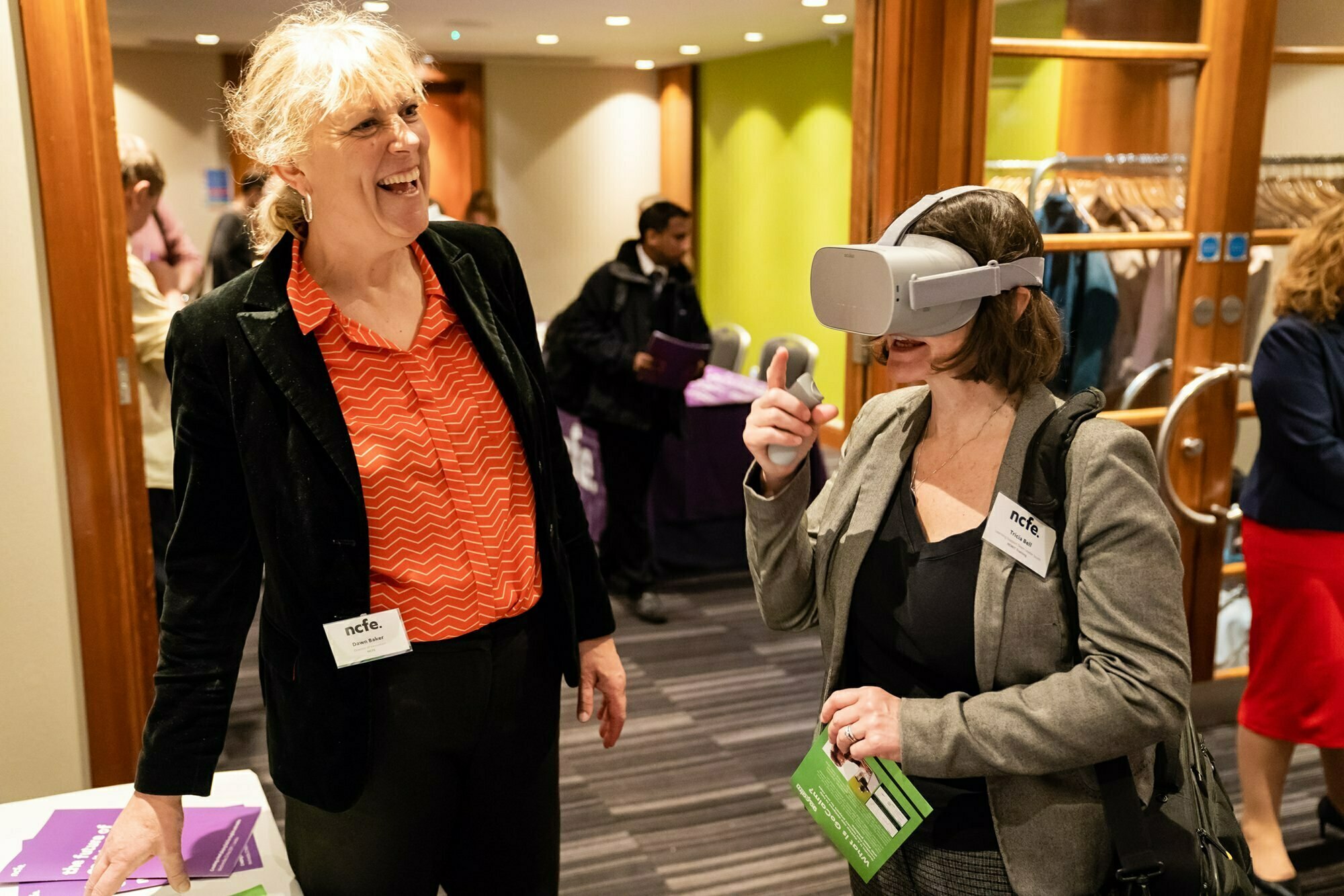 Two women at an event. One, wearing a virtual reality headset, holds informational material and a stylus. The other, standing beside her, smiles while watching. There are attendees and a coat rack in the background. James Gifford-Mead Photography - Event Photographer London