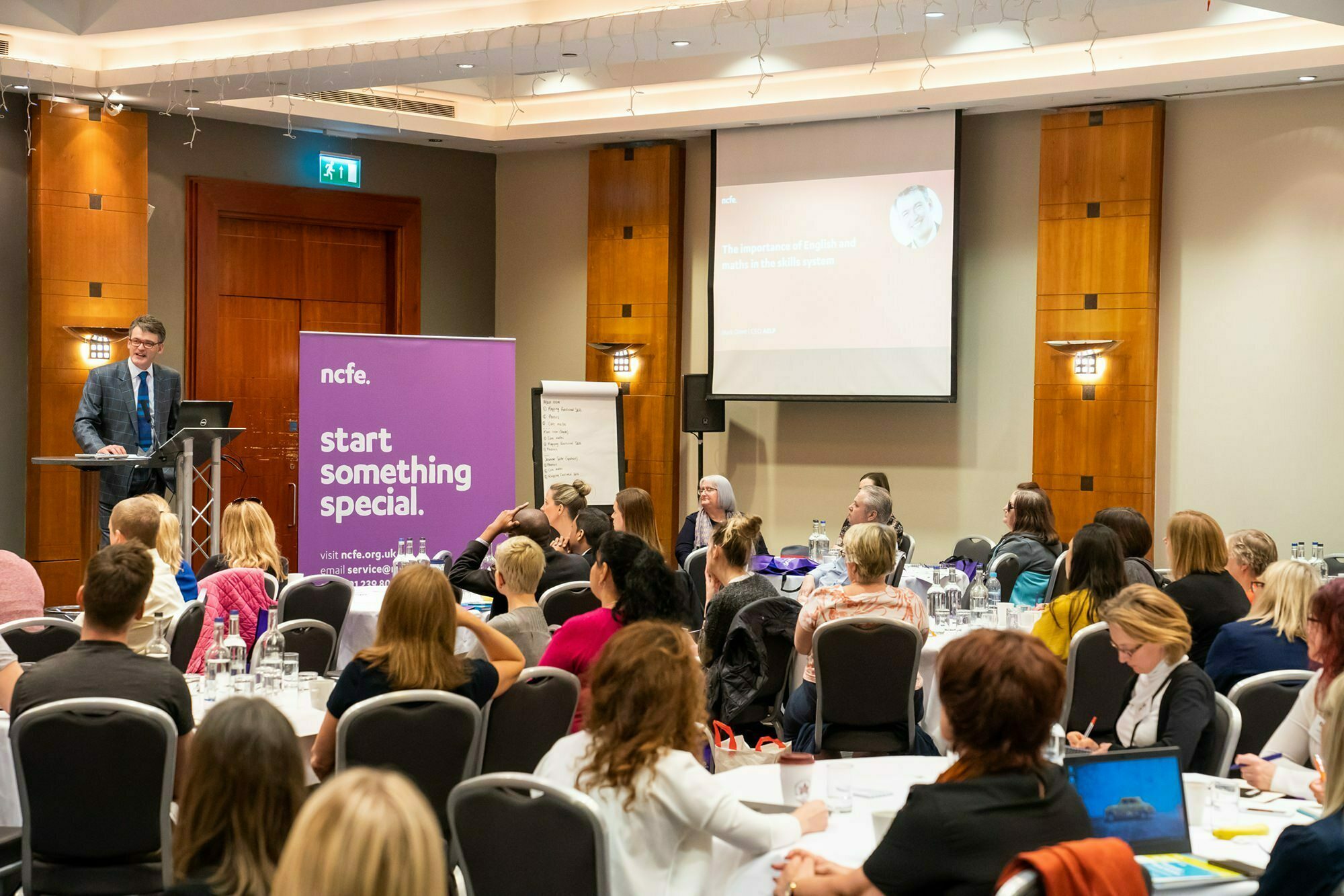 A man in a suit speaks at a podium in front of an audience seated at round tables. A purple banner with the text "start something special" is displayed. A presentation slide with a headshot and text is visible on the wall. James Gifford-Mead Photography - Event Photographer London