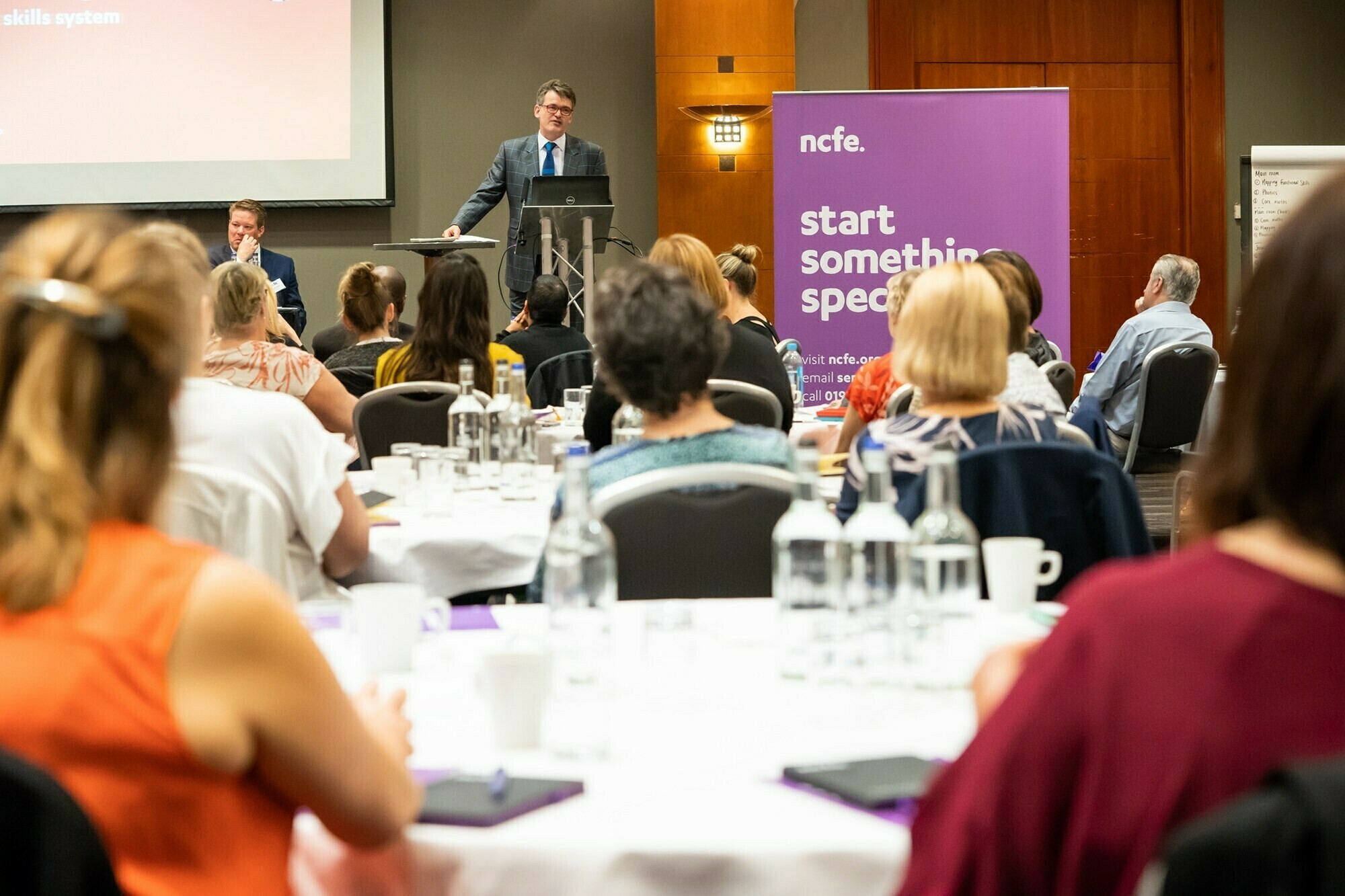 A speaker stands at a podium addressing an audience seated at round tables during a conference. A large purple banner with the text "ncfe. start something special." is visible on the right side. James Gifford-Mead Photography - Event Photographer London