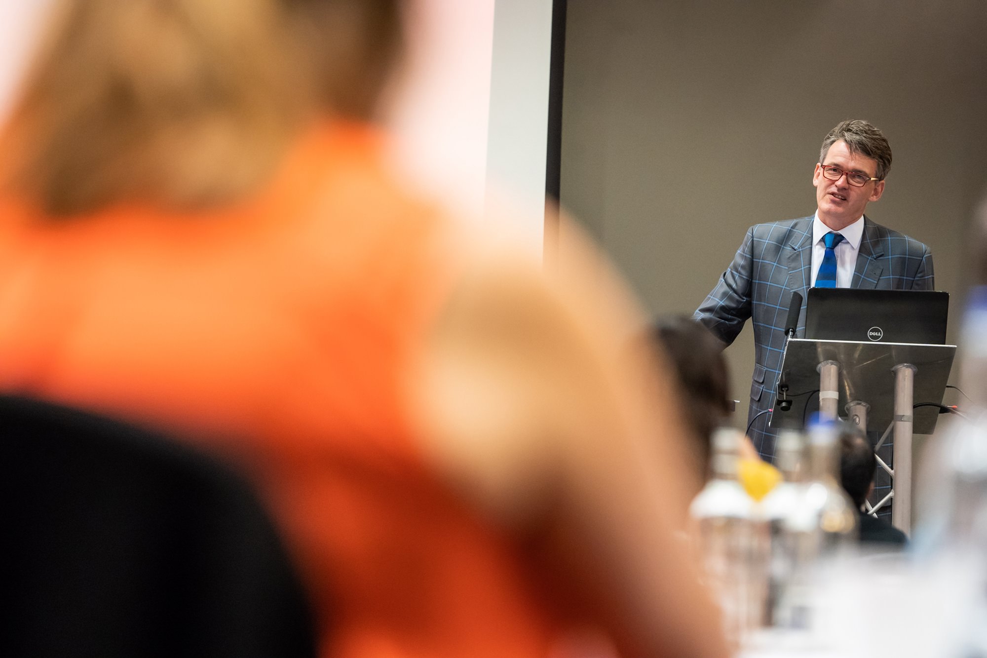 A man in a checkered suit and blue tie speaks at a podium with a laptop, addressing an audience. The foreground is blurred, showing the back of attendees' heads and shoulders. The background is a plain wall. James Gifford-Mead Photography - Event Photographer London