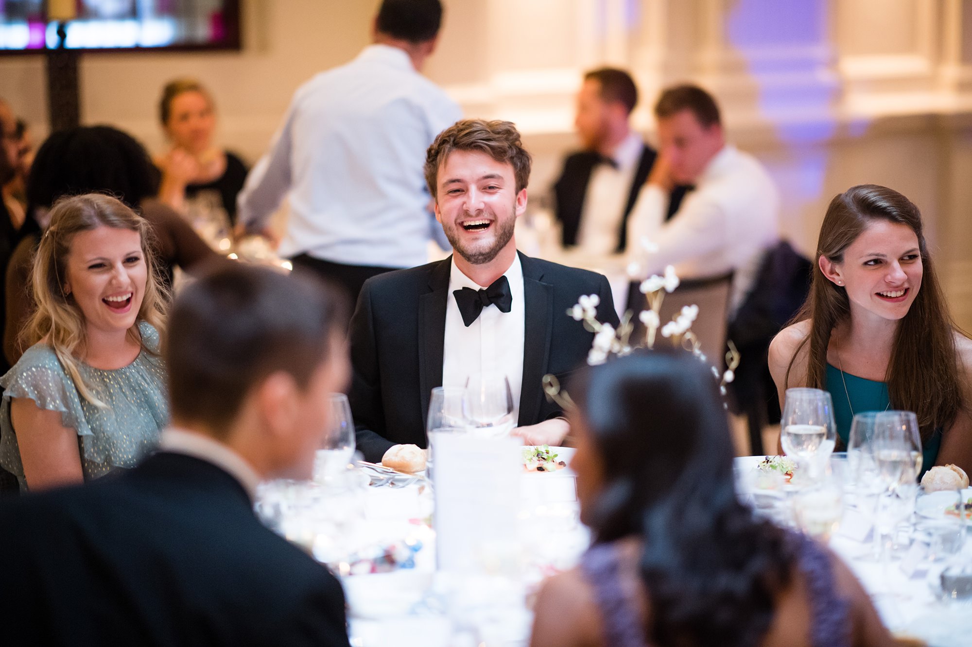 People in formal attire are seated around a round table, smiling and engaging in conversation at an elegant event. The table is set with glassware and decorative arrangements. The background shows more guests seated at similar tables. James Gifford-Mead Photography - Event Photographer London