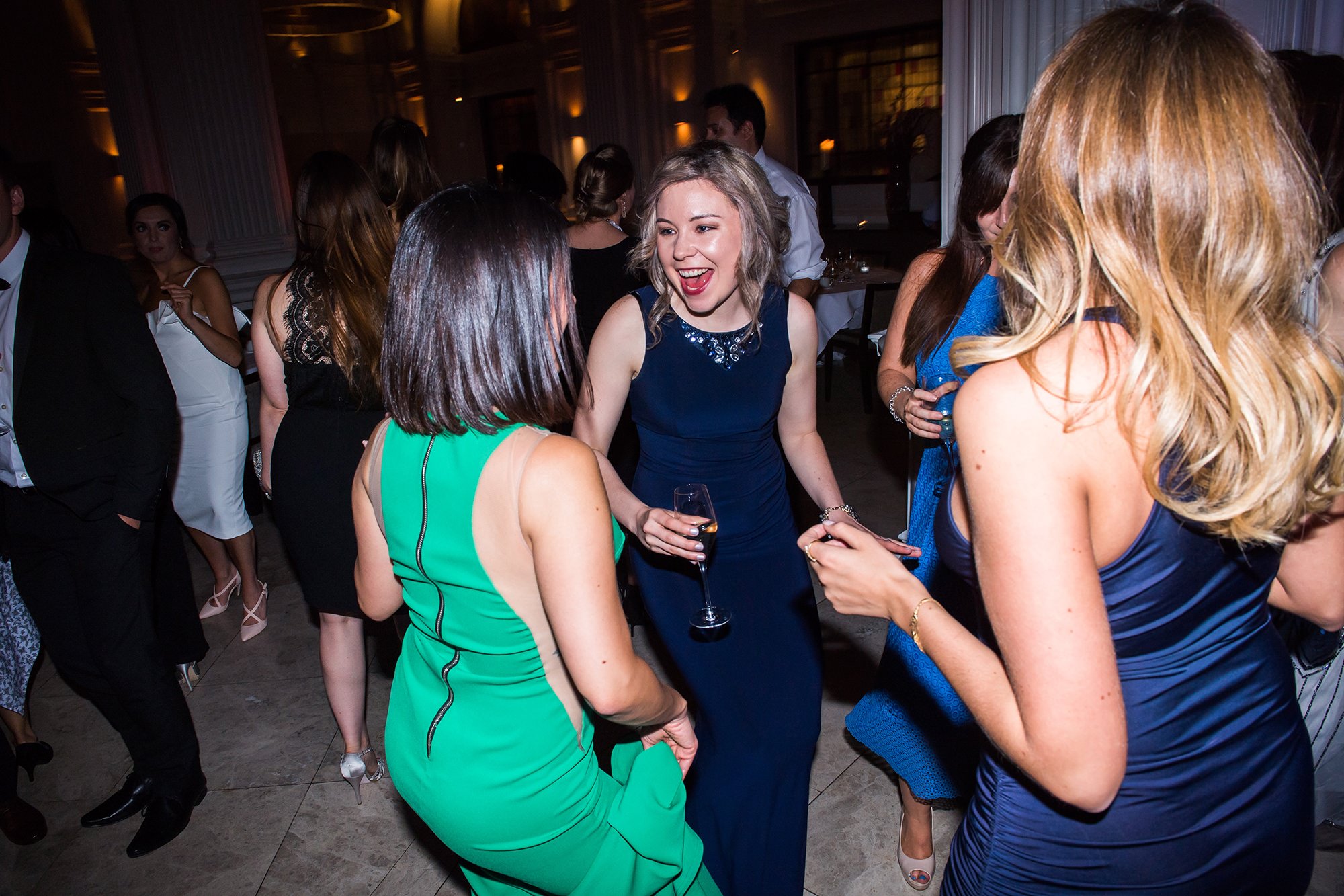 A group of people in formal attire dance energetically at a party. The woman in a blue dress, holding a glass, is smiling and engaging with two women in green and blue dresses. The background is dimly lit, with a warm and festive ambiance. James Gifford-Mead Photography - Event Photographer London
