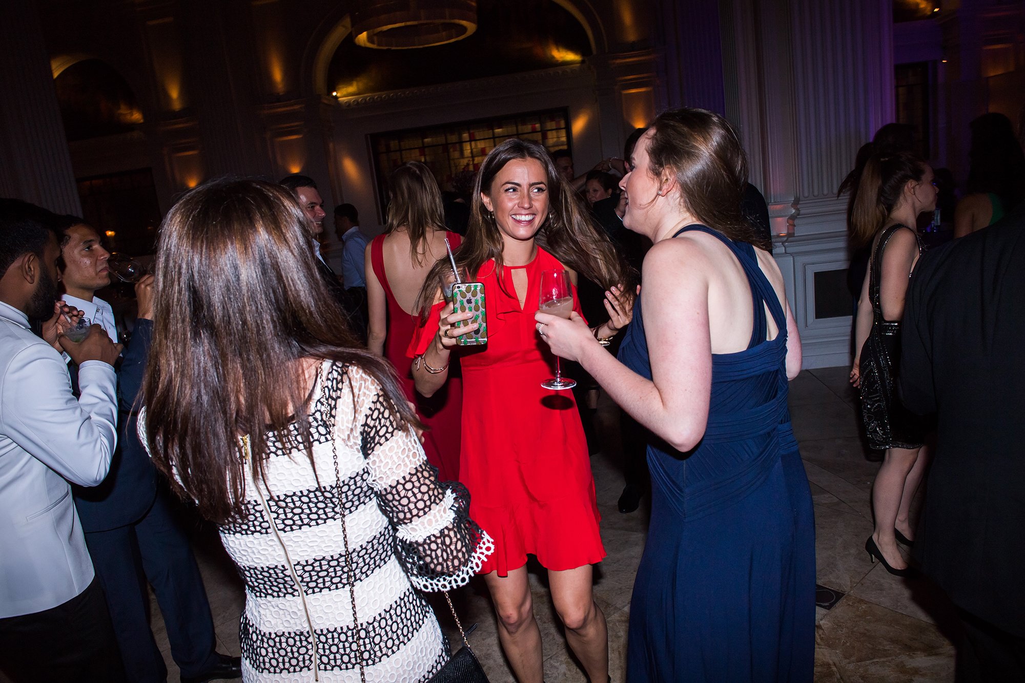 A group of people are dancing at an indoor event. The focus is on a woman in a red dress holding a drink, smiling amid the crowd. Others around her are engaged in conversation and enjoying the lively atmosphere. James Gifford-Mead Photography - Event Photographer London