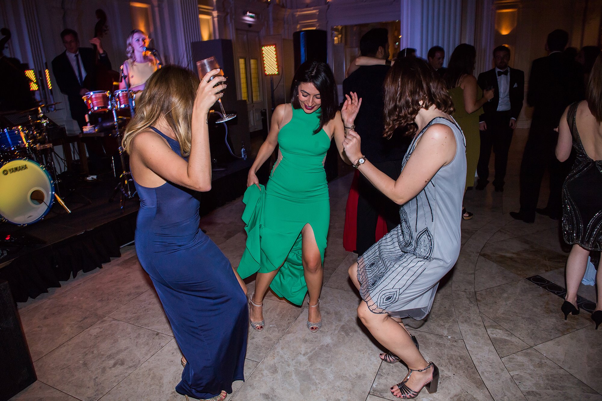 Three women in elegant dresses dance joyfully at a formal event, with a band playing in the background. The venue is decorated with ornate details, and other guests are visible in the background. James Gifford-Mead Photography - Event Photographer London