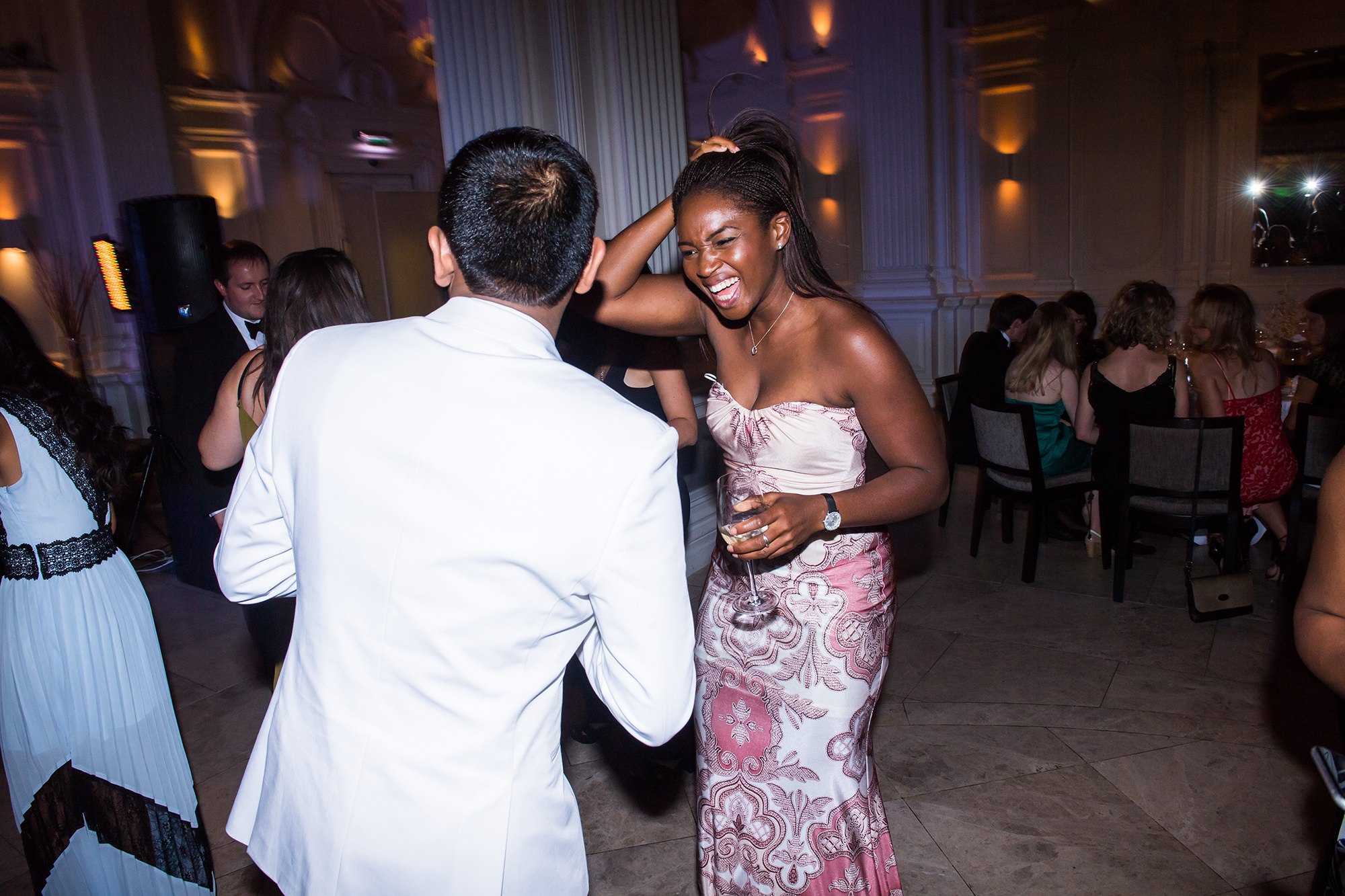 Two people dancing joyfully in a formal setting. The person on the left wears a white suit, while the person on the right wears a patterned strapless dress and holds a drink. The background has elegantly lit architecture and other guests seated at tables. James Gifford-Mead Photography - Event Photographer London