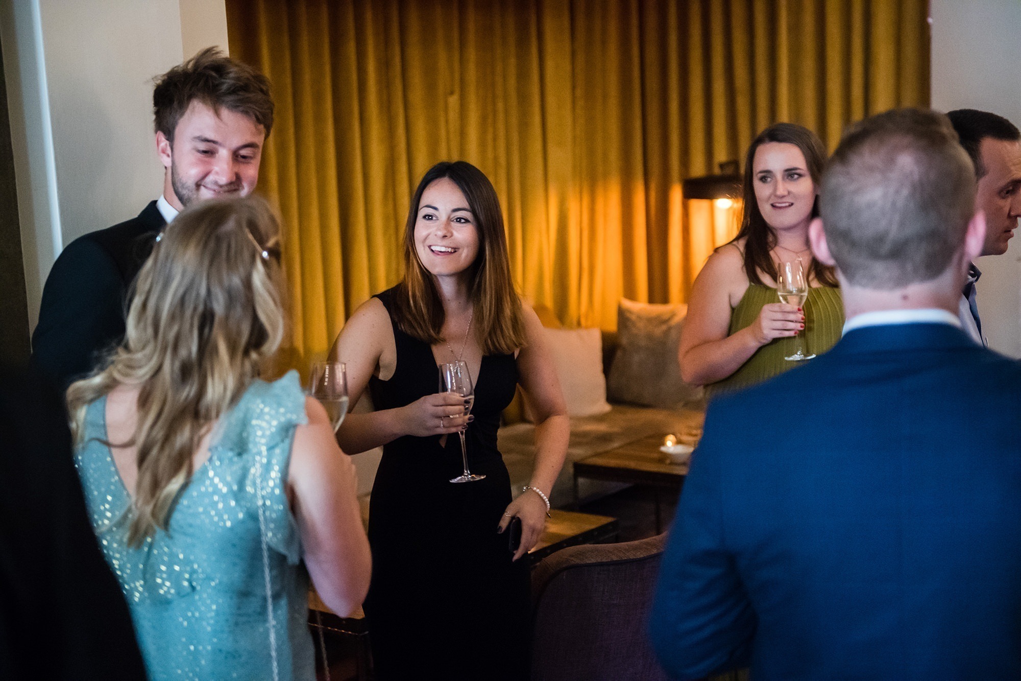 A group of people dressed in formal attire are socializing at a gathering. They are holding glasses with drinks and standing in a room with yellow curtains and ambient lighting. Everyone seems engaged in conversation. James Gifford-Mead Photography - Event Photographer London