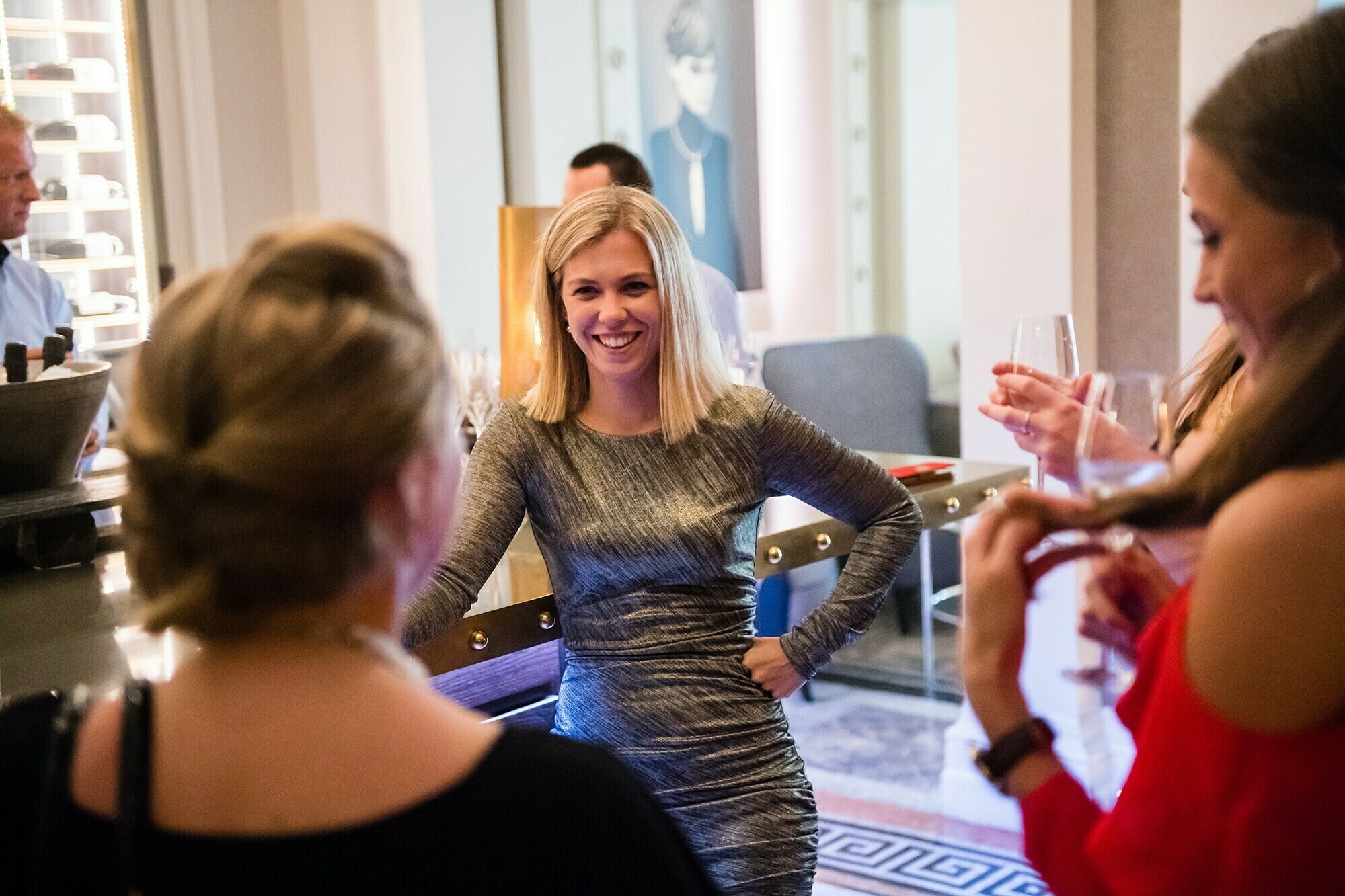 A woman in a gray dress smiles and stands with her hand on her hip in a social setting. She is engaged in conversation with others, who are holding drinks. The atmosphere appears warm and lively. James Gifford-Mead Photography - Event Photographer London