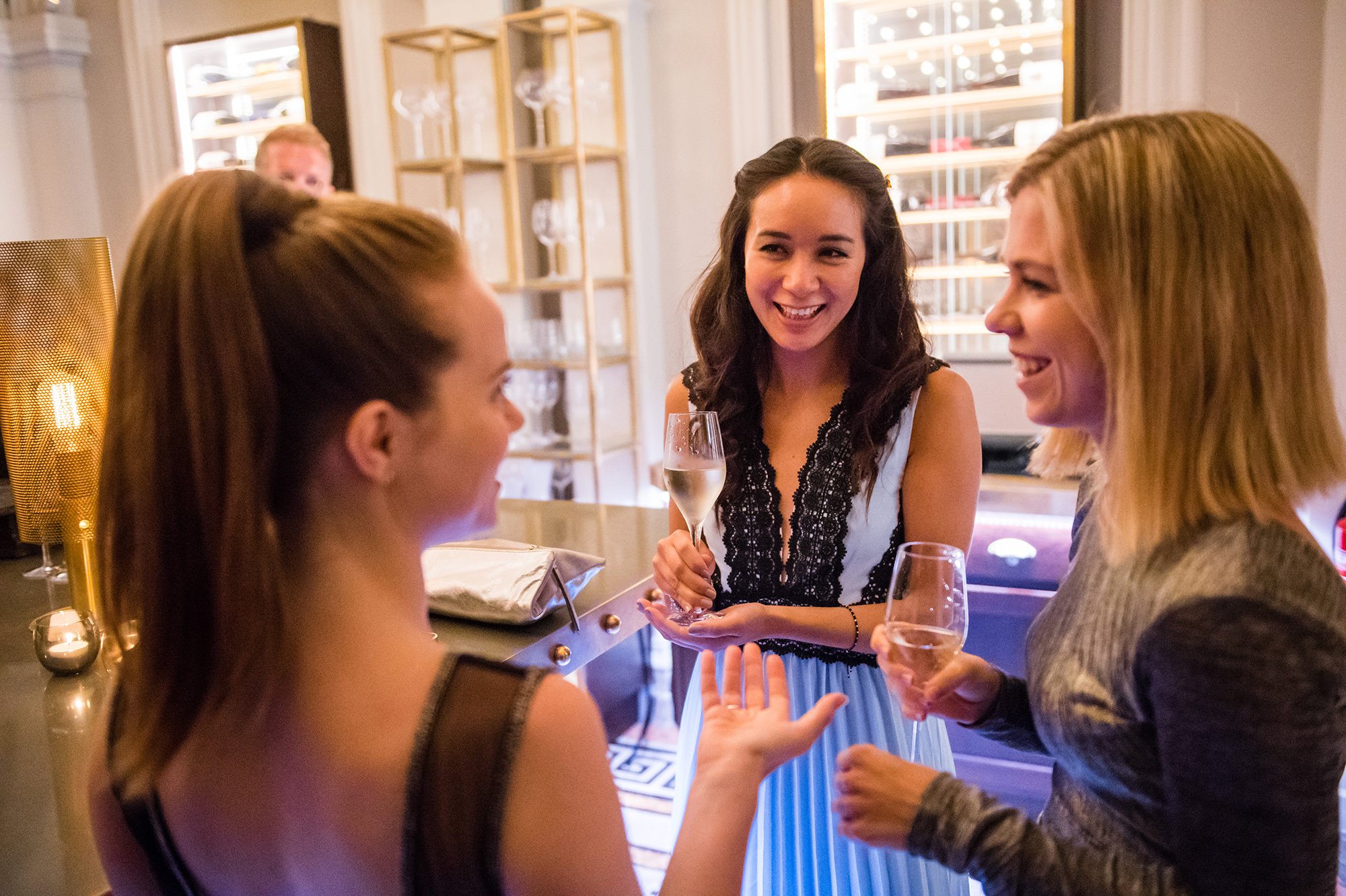 Three women are enjoying a conversation at a social gathering. They are holding glasses of wine and standing near a bar counter in a well-lit room with shelves in the background. They appear to be smiling and engaged in discussion. James Gifford-Mead Photography - Event Photographer London