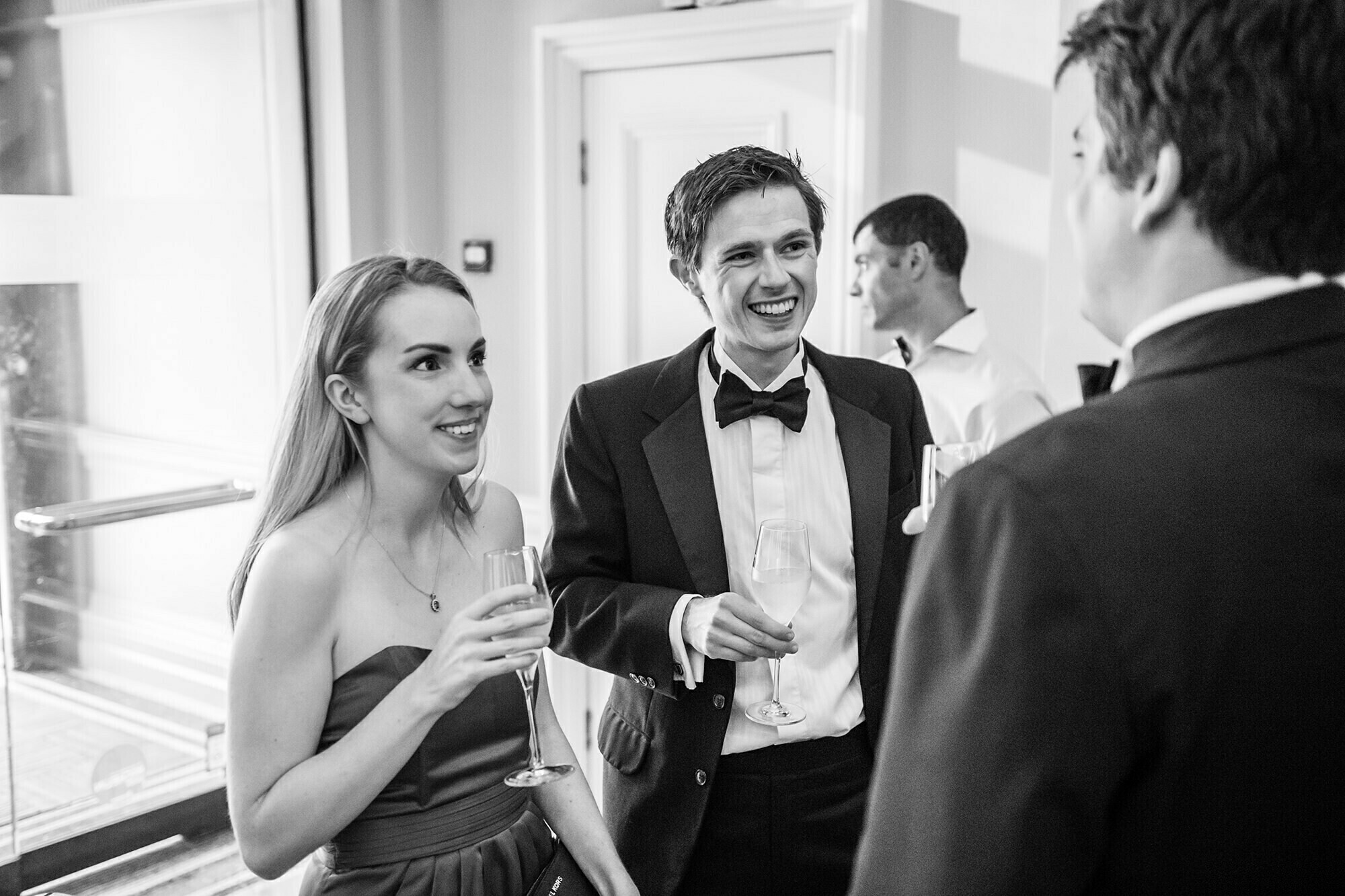 A black-and-white photo of three people in formal attire at a social event. Two are holding glasses and smiling at the third person, who also holds a glass. They stand in a well-lit room with a partially open door in the background. James Gifford-Mead Photography - Event Photographer London