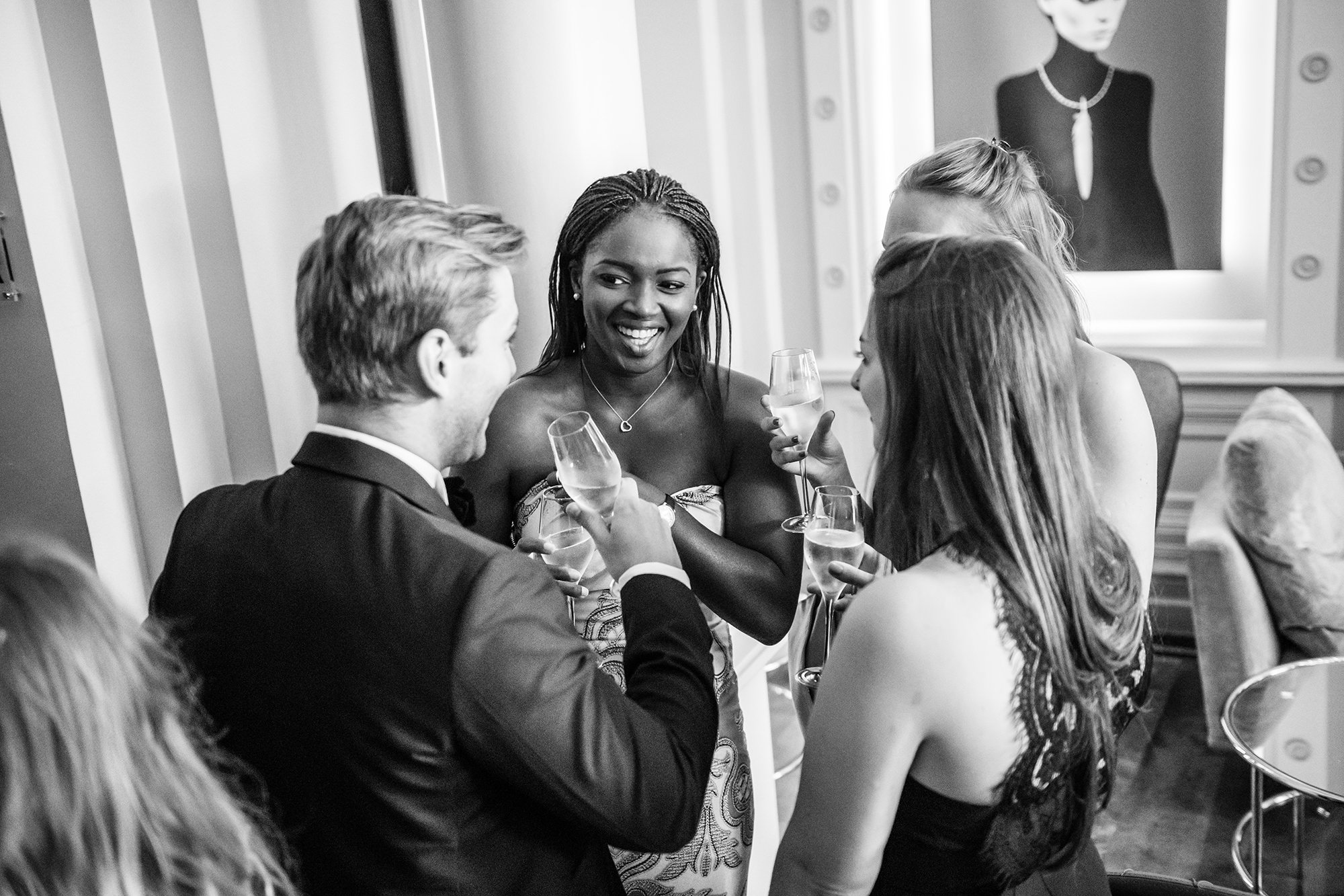 A group of people in formal attire are standing together, holding glasses of champagne and smiling. They're in a stylish room with elegant décor and artwork on the wall. The atmosphere is lively and festive. James Gifford-Mead Photography - Event Photographer London