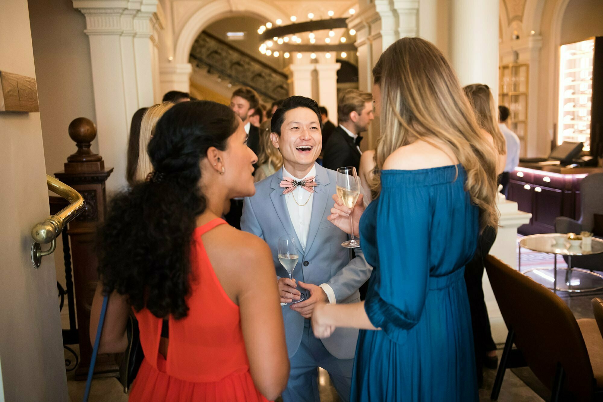 A cheerful group of people socializing at an elegant event. A person in a light blue suit with a bow tie is holding a drink and smiling, while others, dressed in formal attire, stand nearby. The setting is ornate, with arches and warm lighting. James Gifford-Mead Photography - Event Photographer London