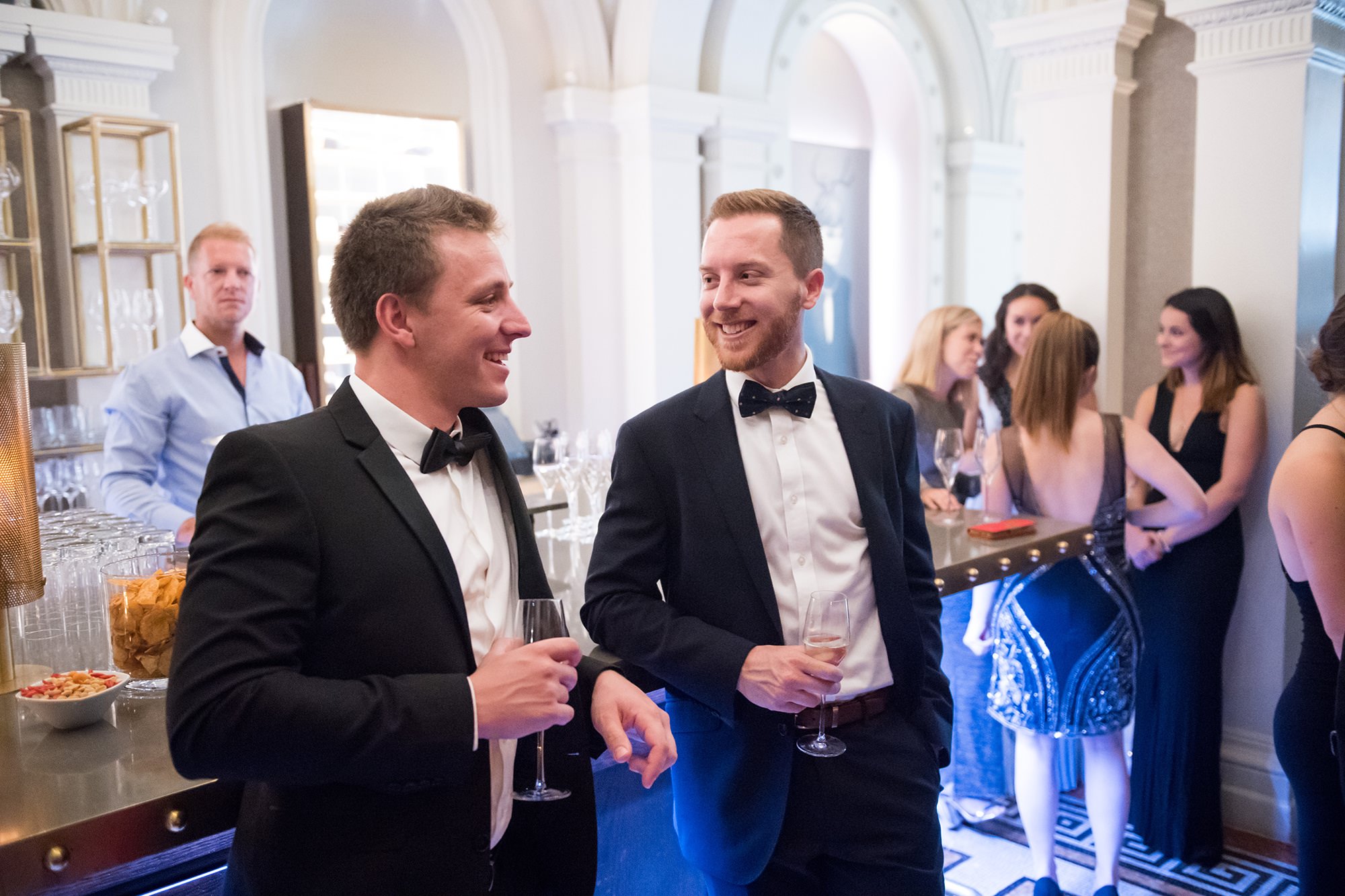 Two men in tuxedos stand at a bar, holding drinks and smiling at each other. In the background, other well-dressed people mingle near a counter with snacks and glasses, against an elegant interior with arched doorways. James Gifford-Mead Photography - Event Photographer London
